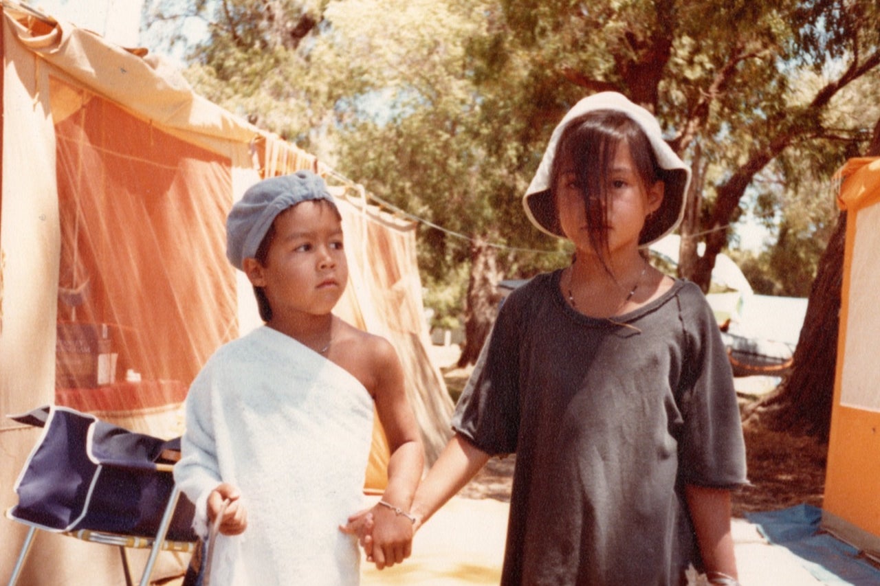 Two young children holding hands outside, with a tent and trees in the background.