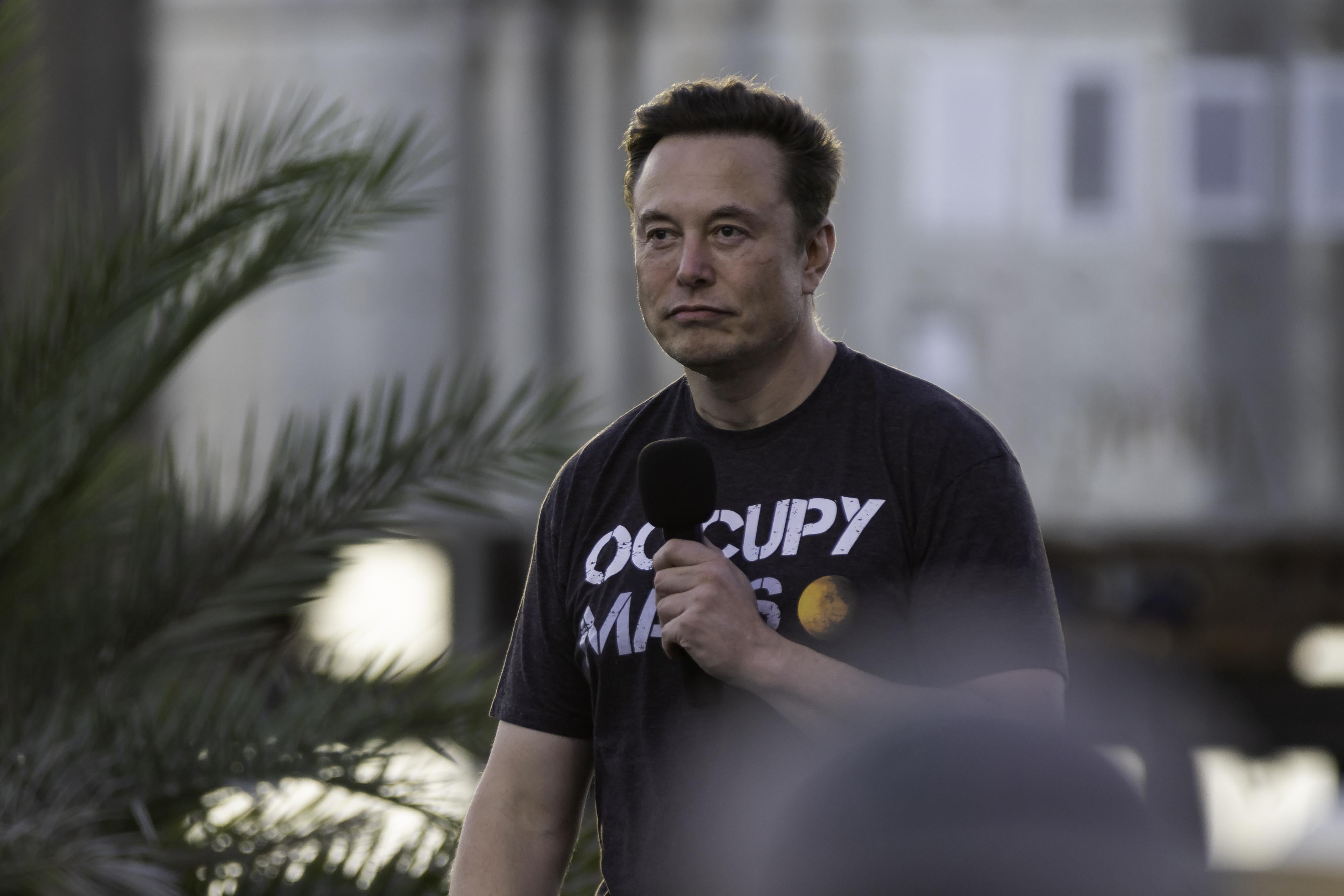 Elon Musk frowns while holding a microphone. He wears a T-shirt that says, "OCCUPY MARS."