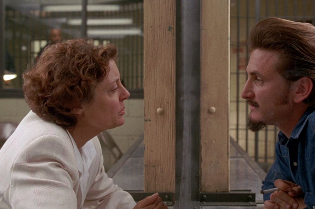 The profile of Susan Sarandon faces right looking at the left facing profile of Sean Penn behind a pane of glass as he's dressed as a prisoner.