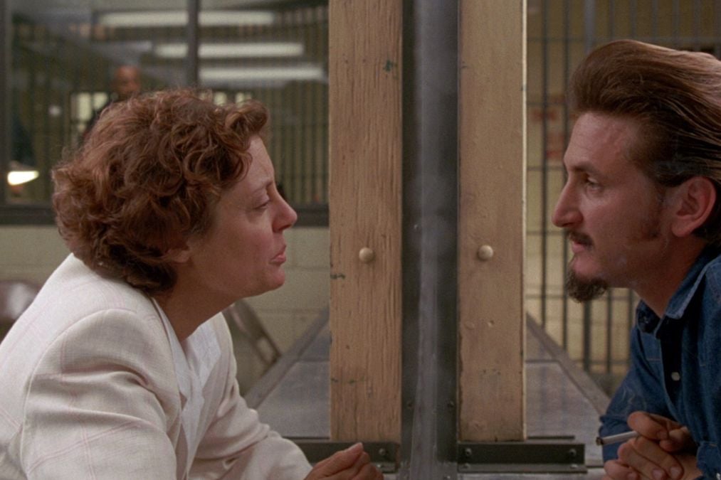 The profile of Susan Sarandon faces right looking at the left facing profile of Sean Penn behind a pane of glass as he's dressed as a prisoner. 