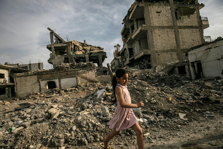 A girl in a pink dress walks in front of rubble and destroyed buildings.