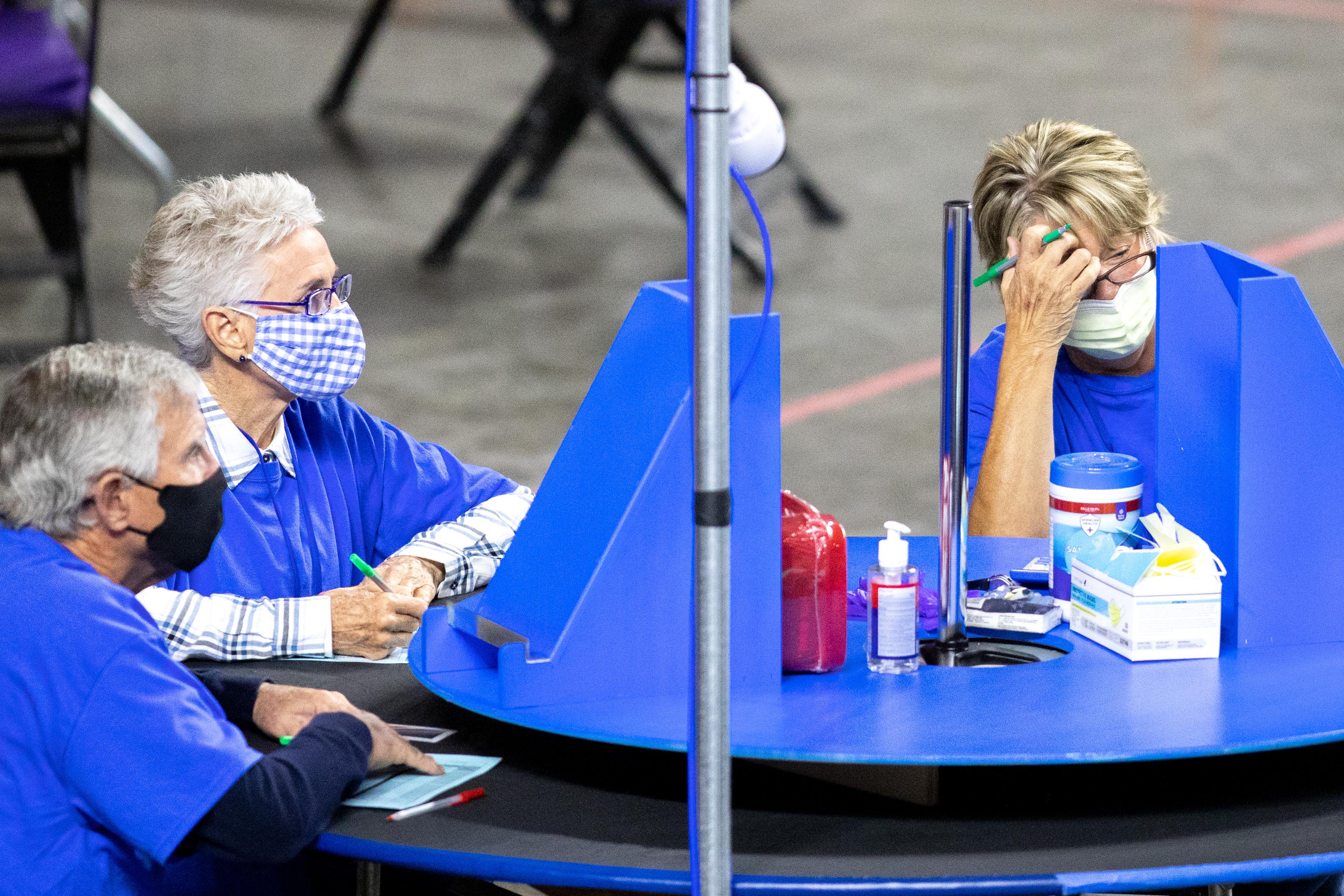 A woman leans her head into her hand in apparent frustration as she sits in front of a blue Lazy Susan on a table. Two other older people sit at the table.
