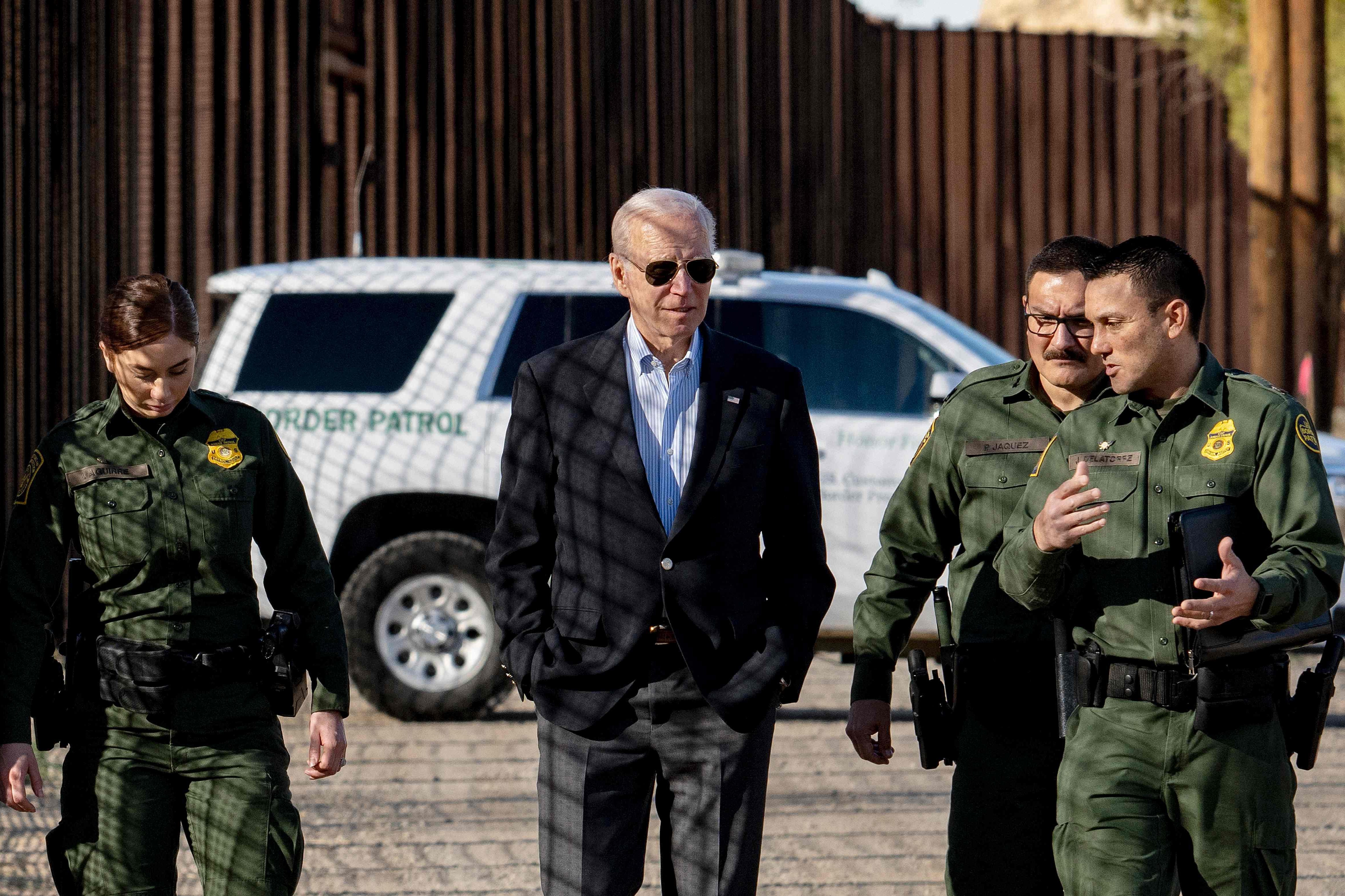 Biden and CBP officers walk together with a Border Patrol SUV and a tall border fence looming and receding behind them