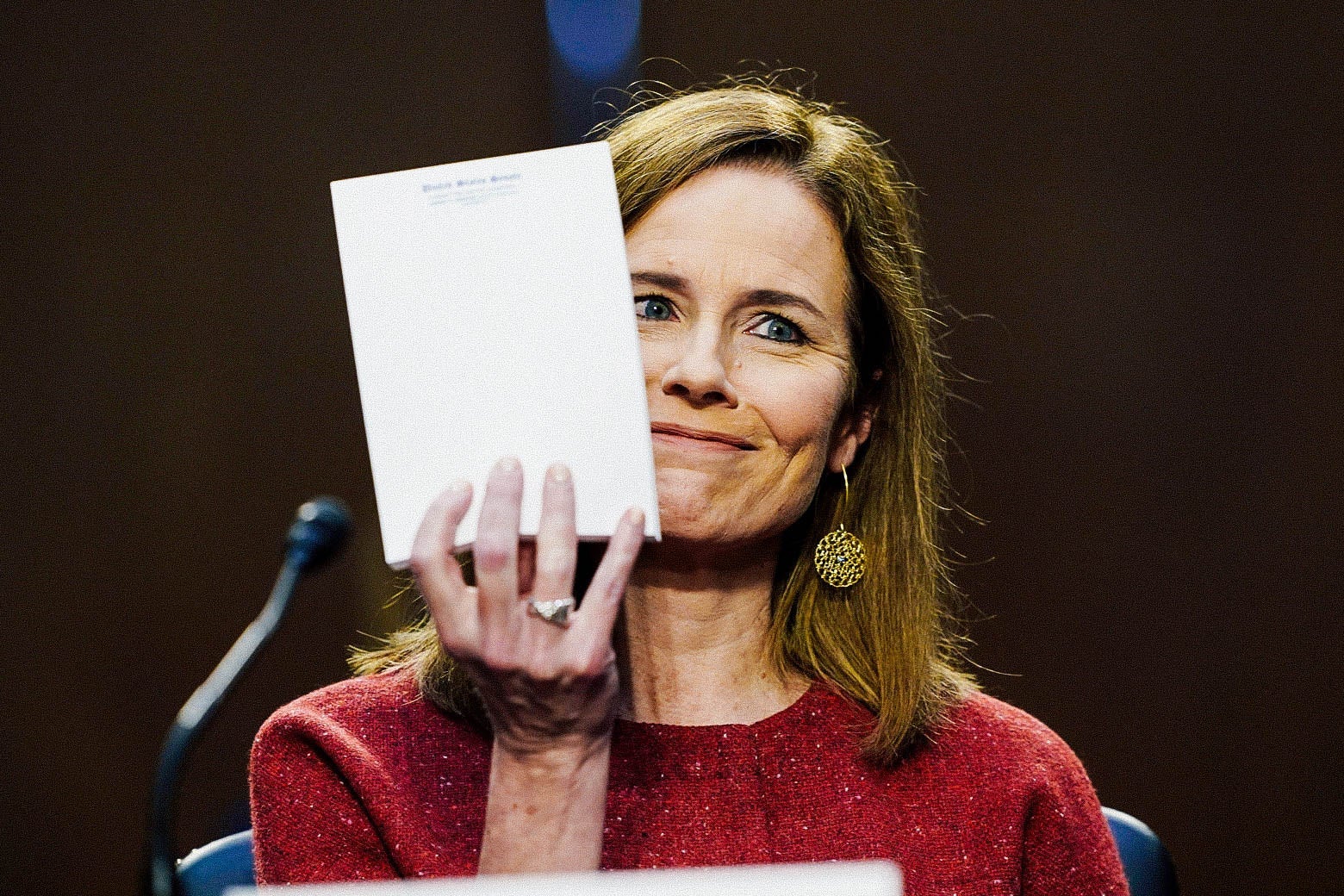 A white woman in a red sweater holds up a blank notepad and smirks.