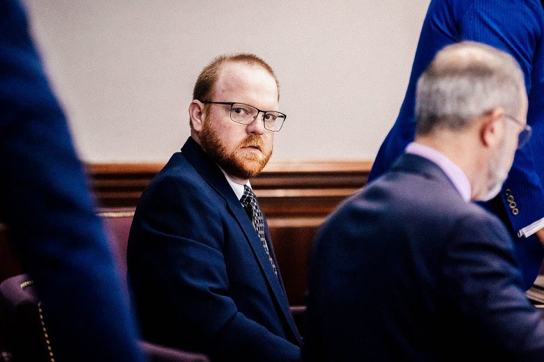 A white man with a beard wearing glasses and a navy suit looks back from a table in a courtroom.