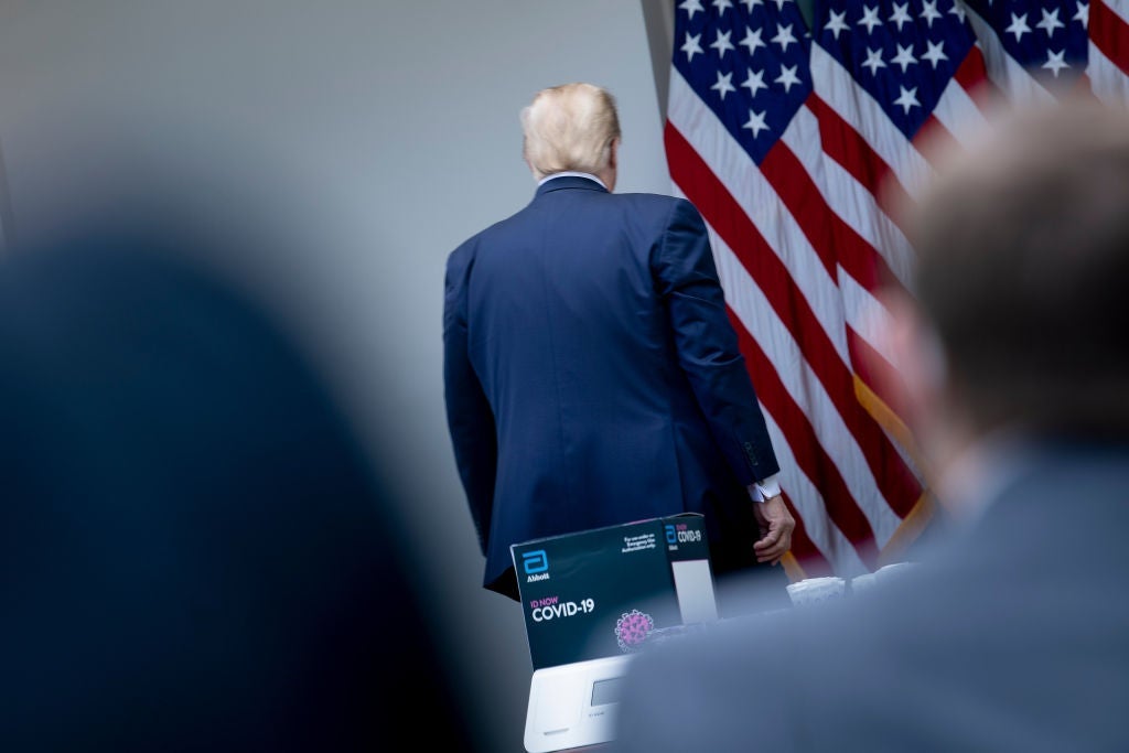 Trump's back is seen in focus from between the blurry shapes of two individuals watching him leave. 