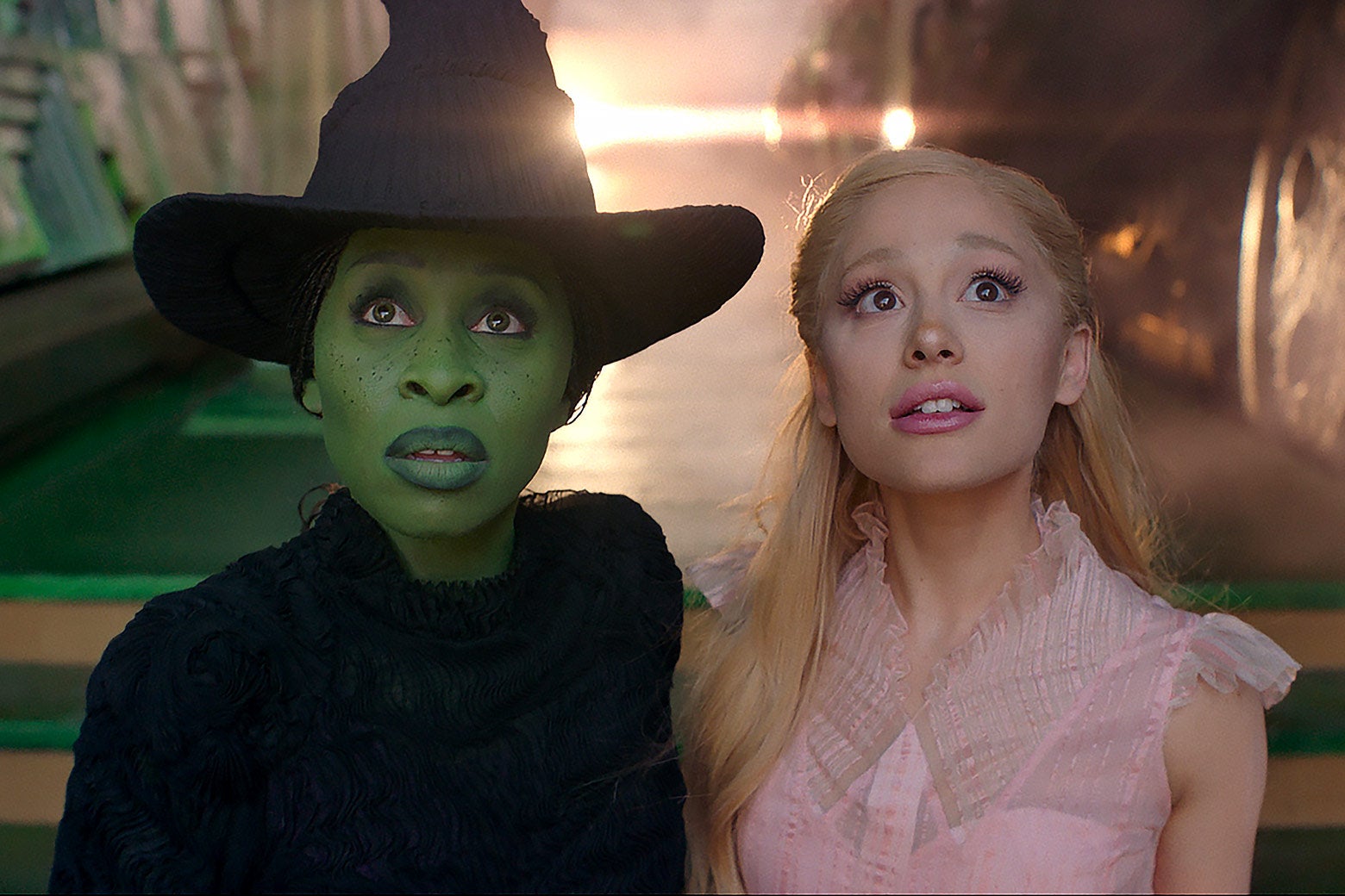 The two actresses look up in awe. Erivo has the familiar look of a pointy black hat, a black dress, and green skin, while Grande is dressed all in bubblegum pink.