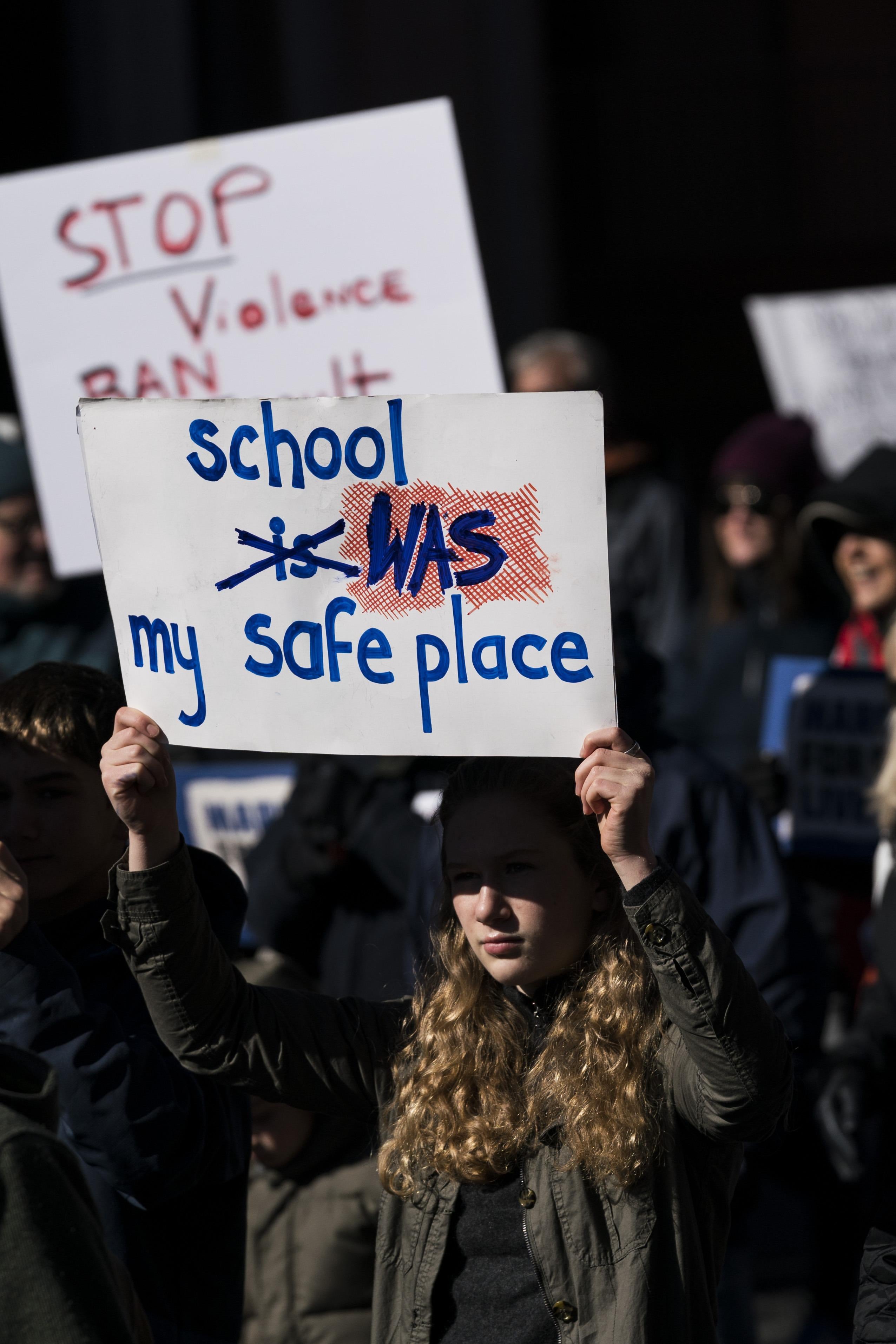 A young woman holds up a sign that reads, "School is [crossed out] was my safe place."