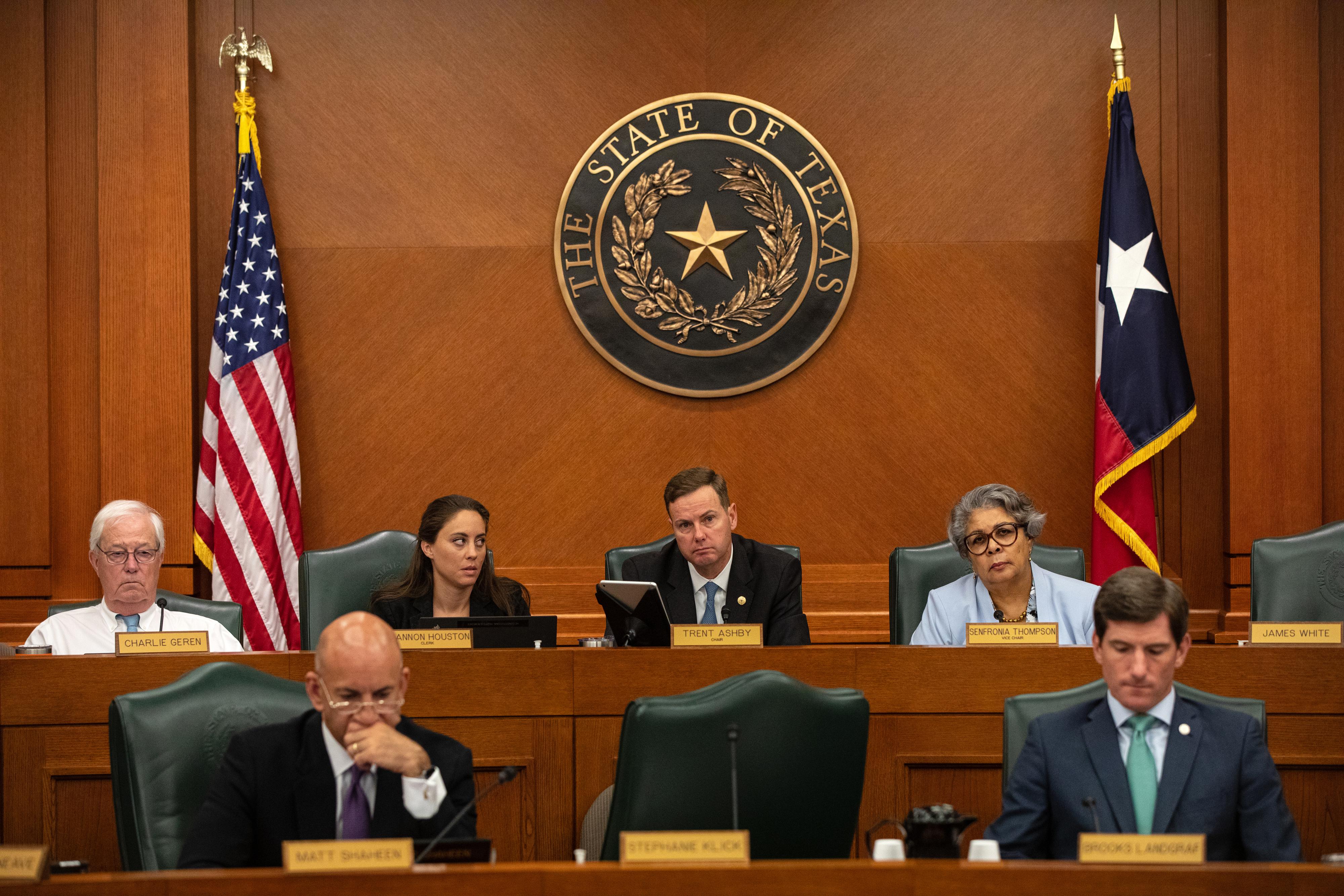 State legislators sit in two raised rows in a committee hearing room in front of American and Texas flags, with the state seal centered on the wall behind them.