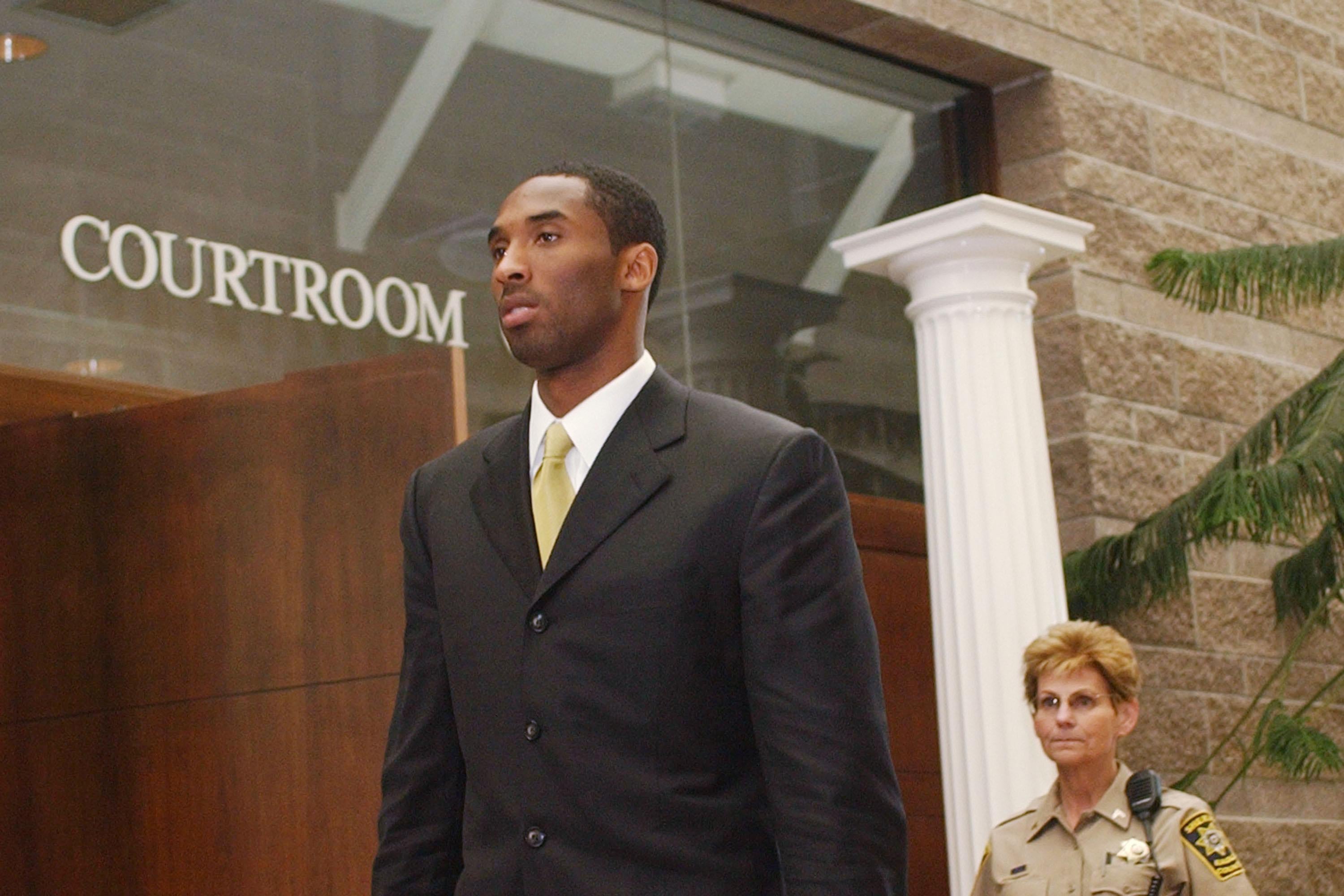 Kobe Bryant, in a suit, walks under a wall that says "Courtroom" as a woman in a law enforcement uniform stands behind him.