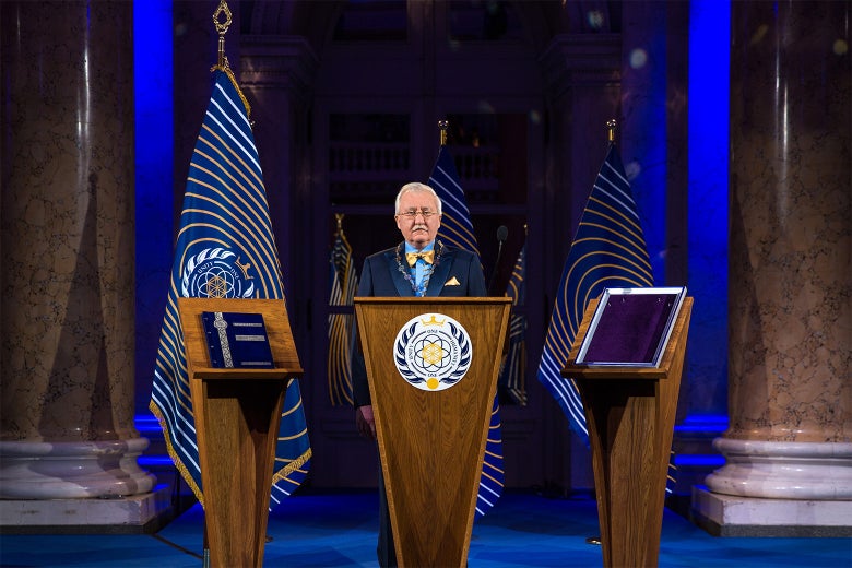 Asgardia President Igor Ashurbeyli stands behind a podium flanked by the flag of Asgardia.