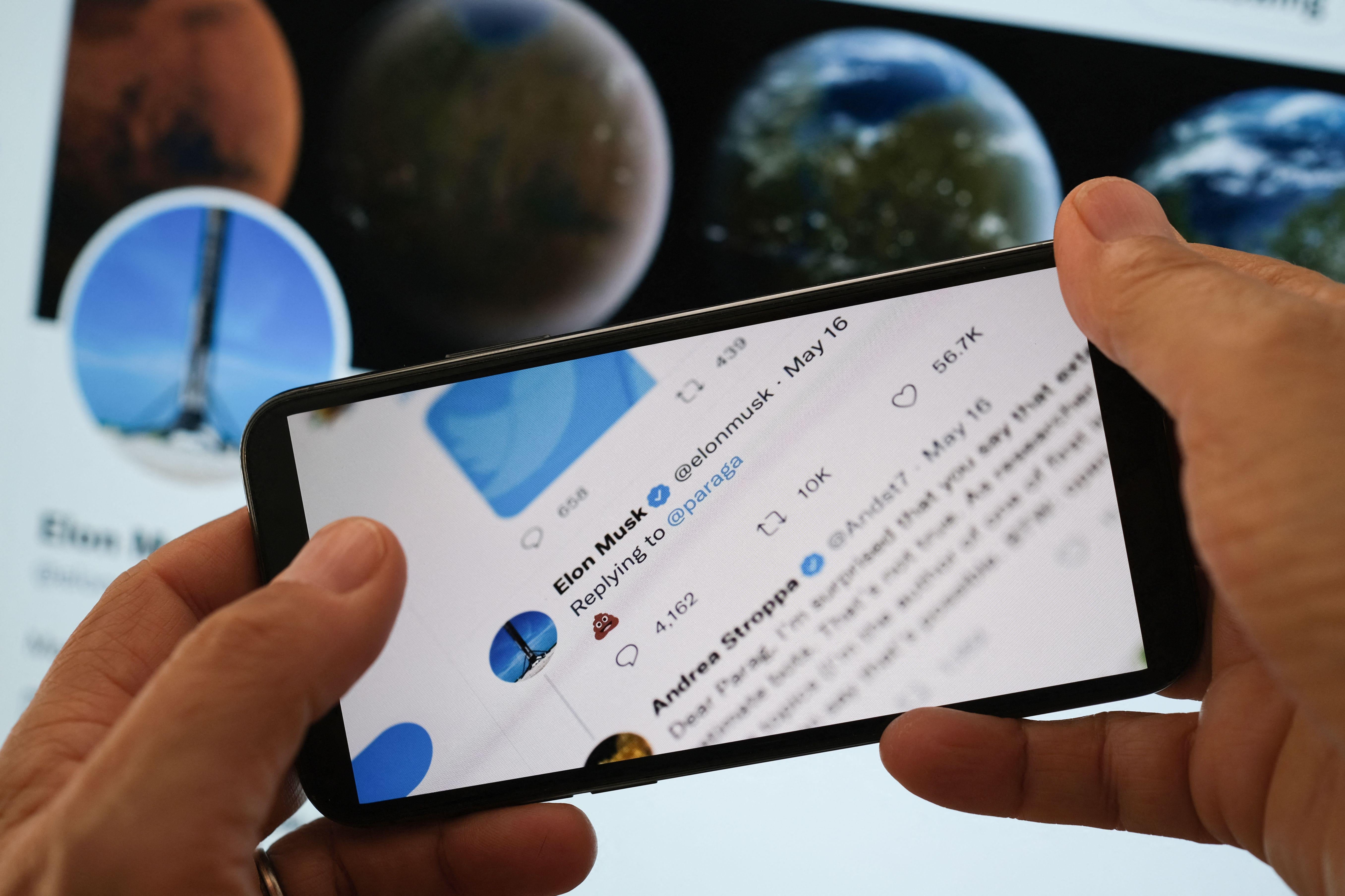 Hands holding a smartphone—which is displaying Elon Musk's tweet of a poop emoji in response to Twitter's CEO—in front of a larger screen displaying Musk's Twitter account.