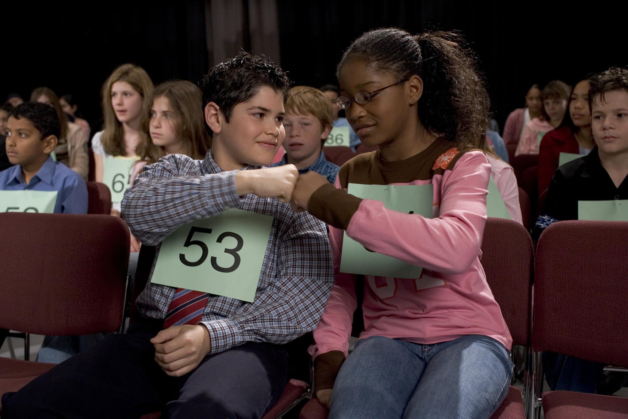 A young Keke Palmer fist bumps a young boy she's sitting next two. Both of them are spelling bee contestants sitting in an auditorium with other contestants behind them.