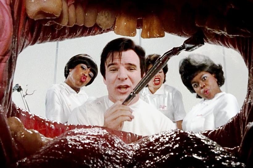 The view from inside a mouth: a dark-haired Steve Martin, dressed as a dentist, framed by a tongue and teeth. Behind him, three women dressed as nurses.