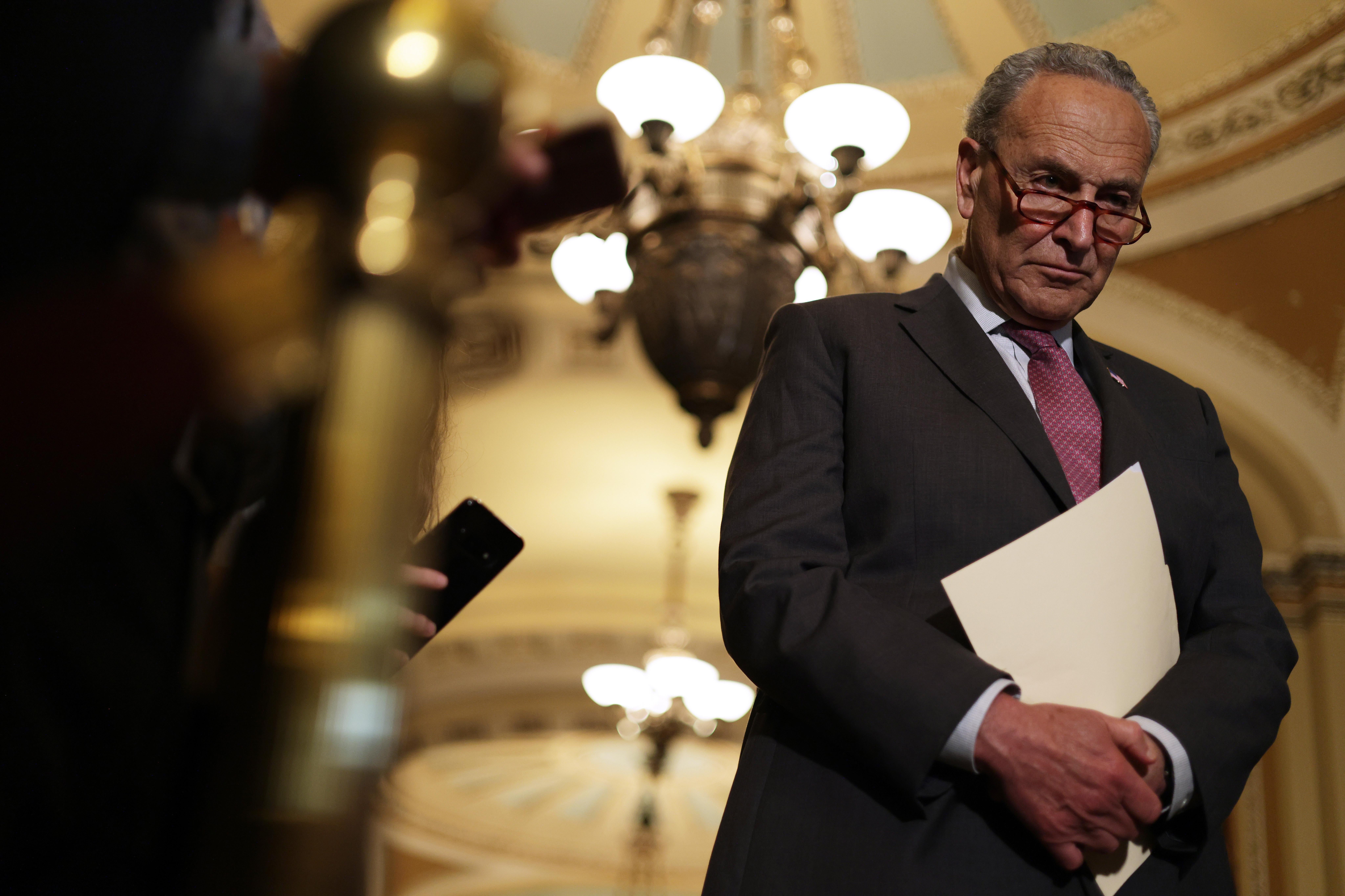 Low-angle shot of Schumer frowning as he stands holding a manila folder to his chest