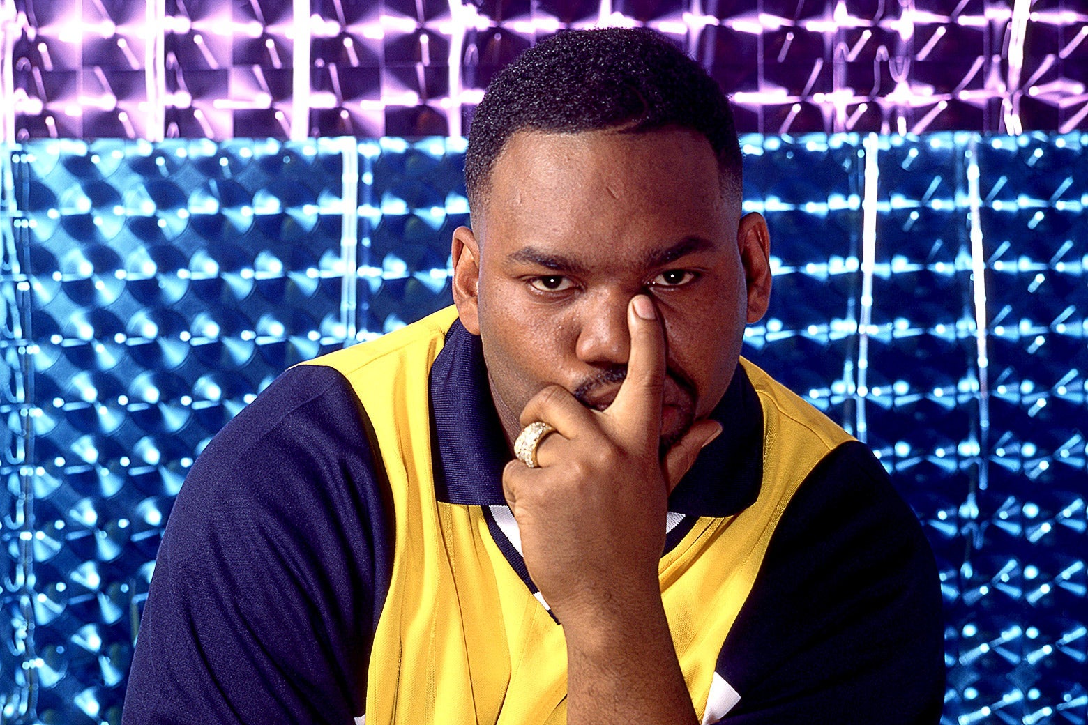 Raekwon of the Wu-Tang Clan posing in front of a sparkly backdrop in a yellow and navy blue shirt.