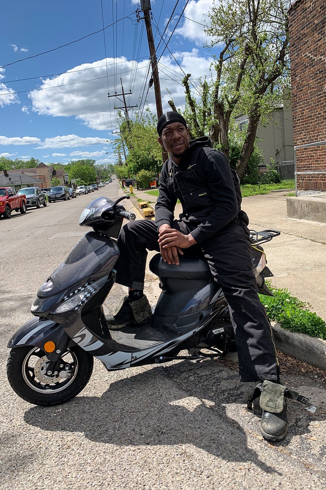 A Black man in a black do-rag, a black sweatshirt, and black overalls sits on a motorized scooter on the side of a street.