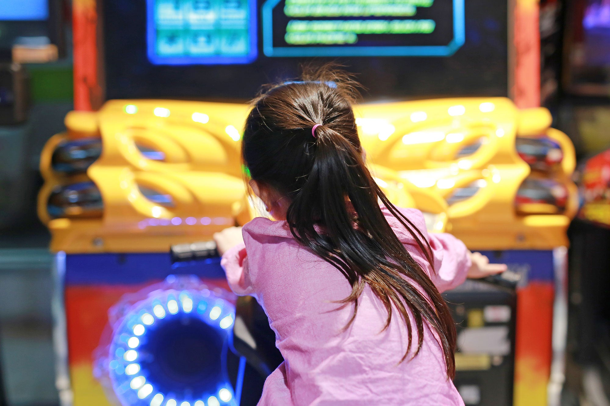 A ponytailed child in a pink sweatshirt faces an arcade game.