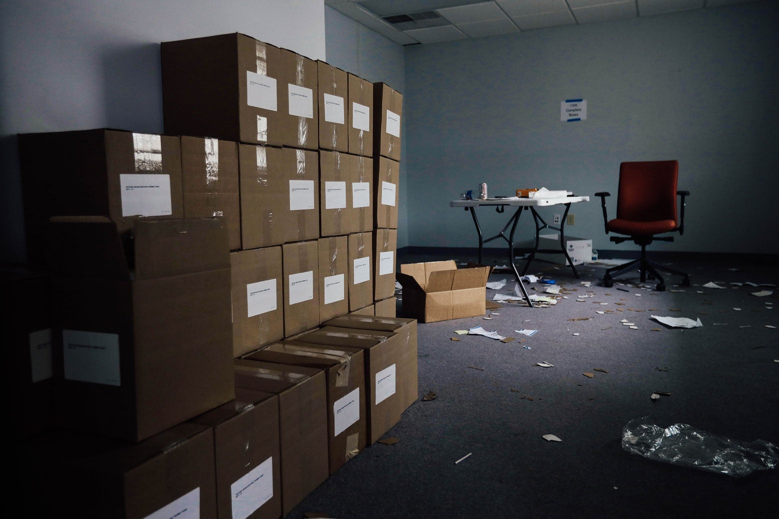 Boxes are stacked in room. In the back, a desk and chair stand, with pieces of paper scattered around.
