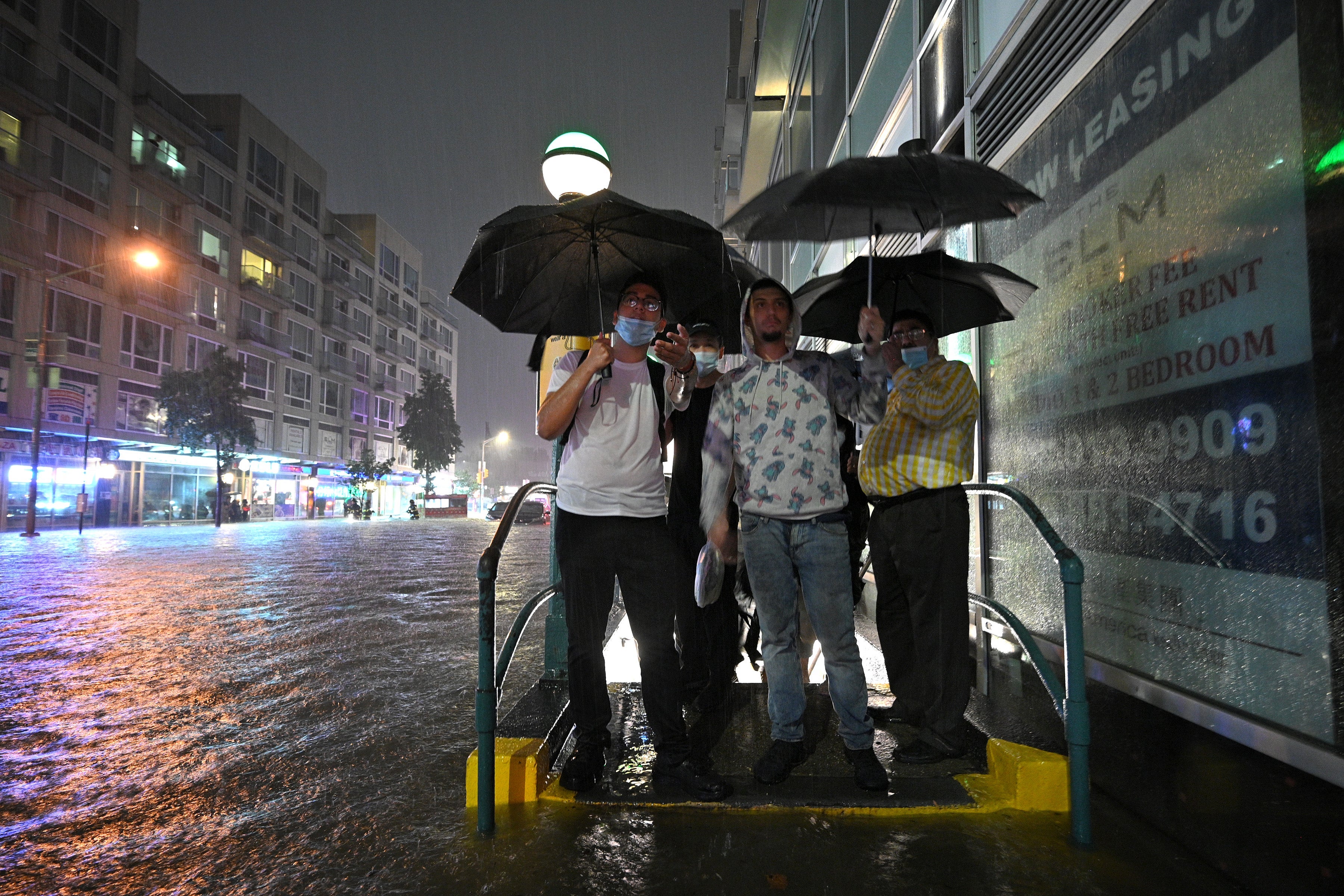 People holding umbrellas stand huddled at a subway entrance as water fills the street and sidewalk.