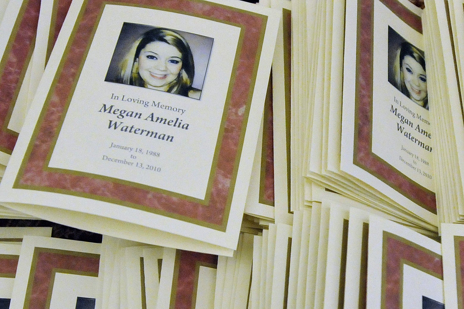 A stack of funeral programs featuring a photo of a young woman and the words "In Loving Memory: Megan Amelia Waterman."