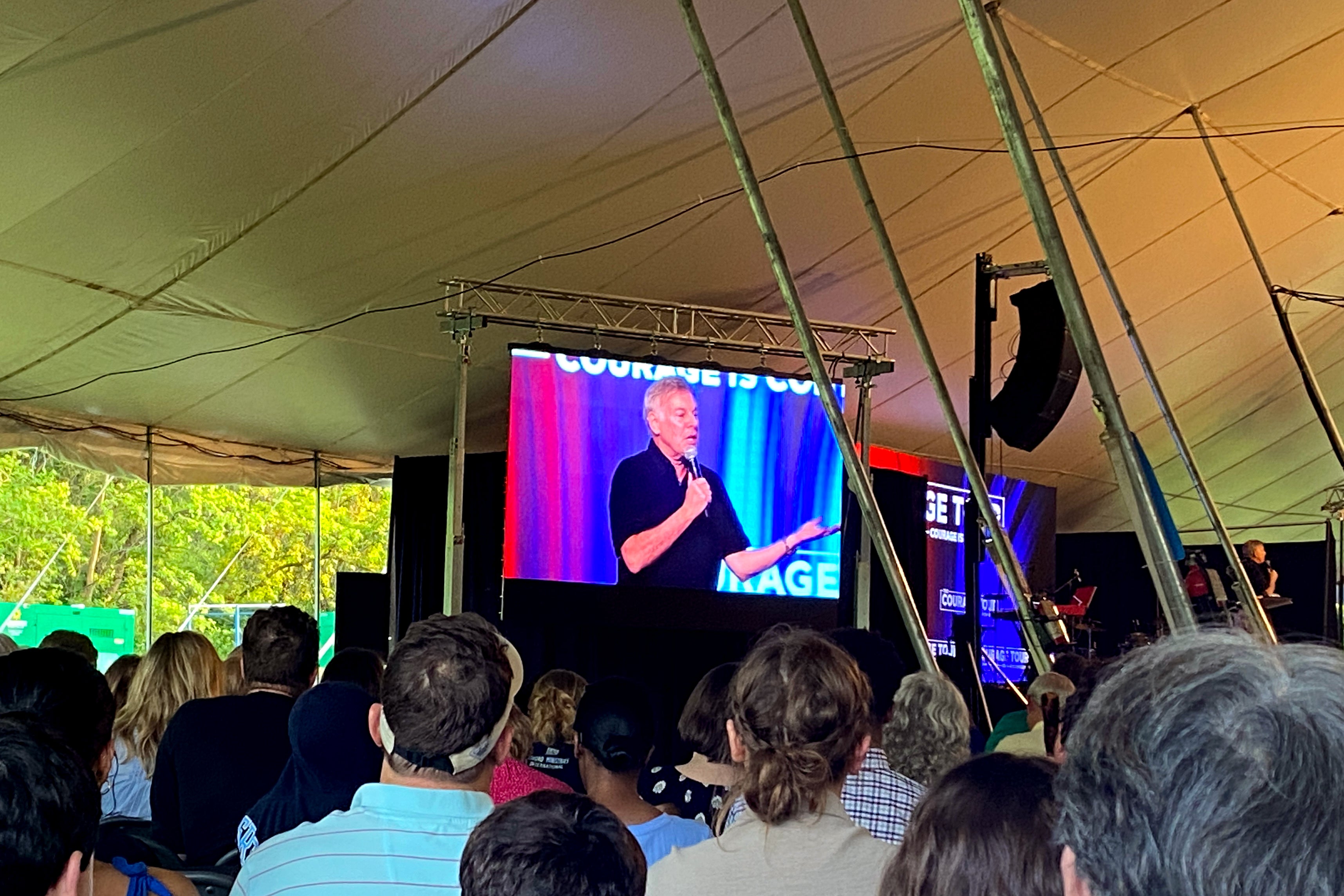 A white man with gray hair speaks on a screen at a tent revival.