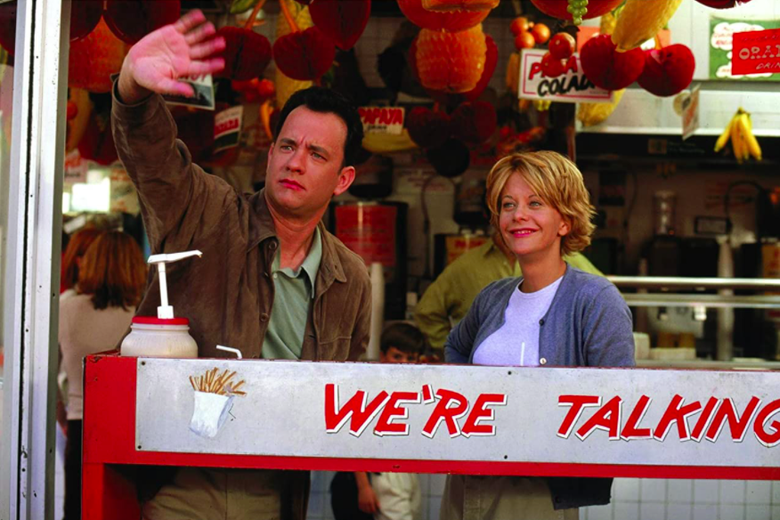 Tom Hanks waves to someone off camera as Meg Ryan stands smiling next to him.