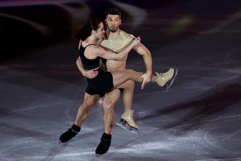 An apparently naked man in ice skates carrying a woman in a little black dress and ice skates