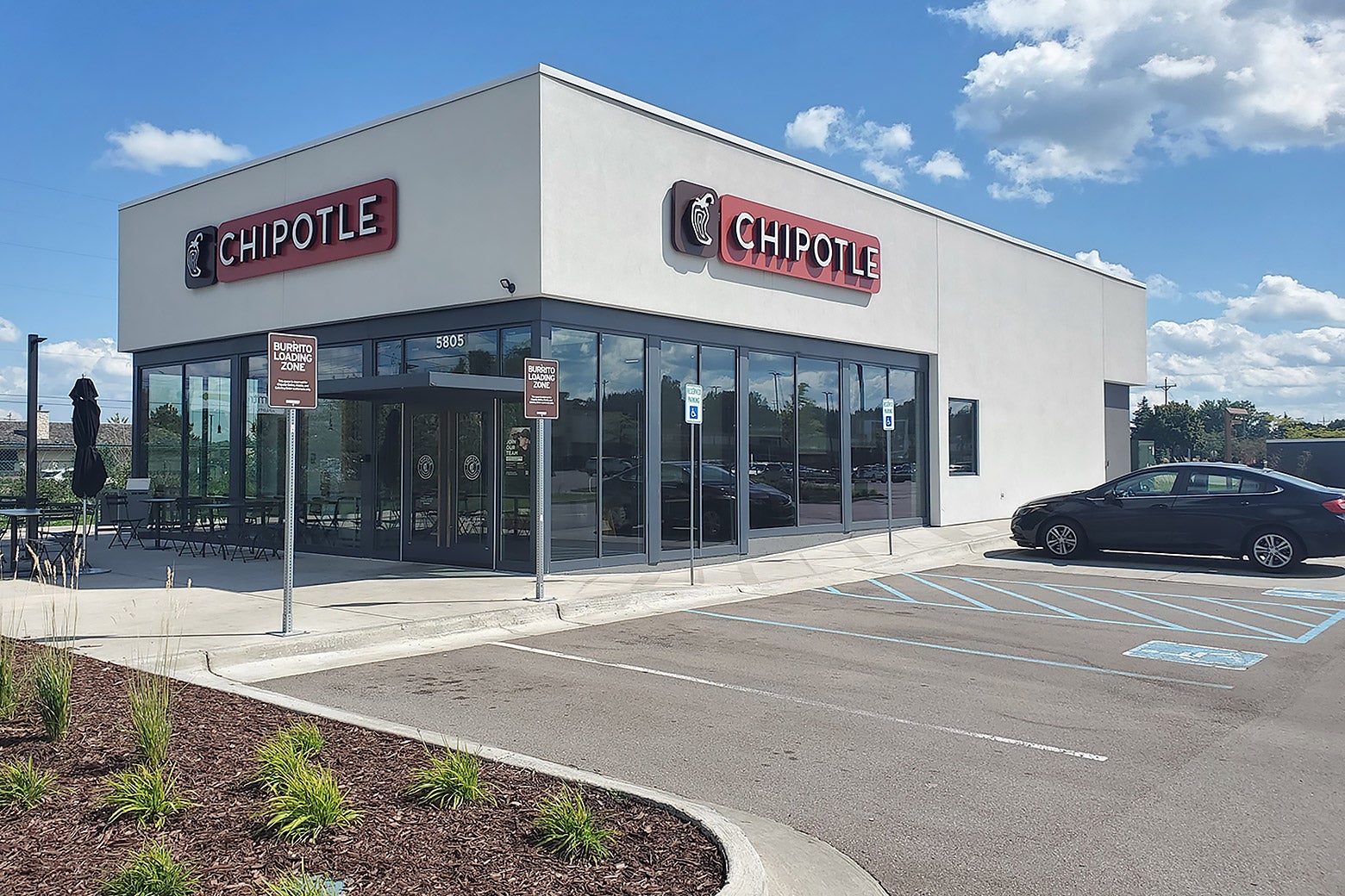 A parking lot and a modern-looking building with large glass windows in the front that says CHIPOTLE on two sides.