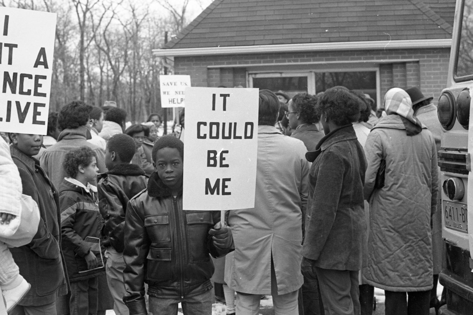 A black-and-white photo shows a black child carrying a serious expression and a sign that reads "It could be me." He is standing facing away from a crowd that is staring at a house.