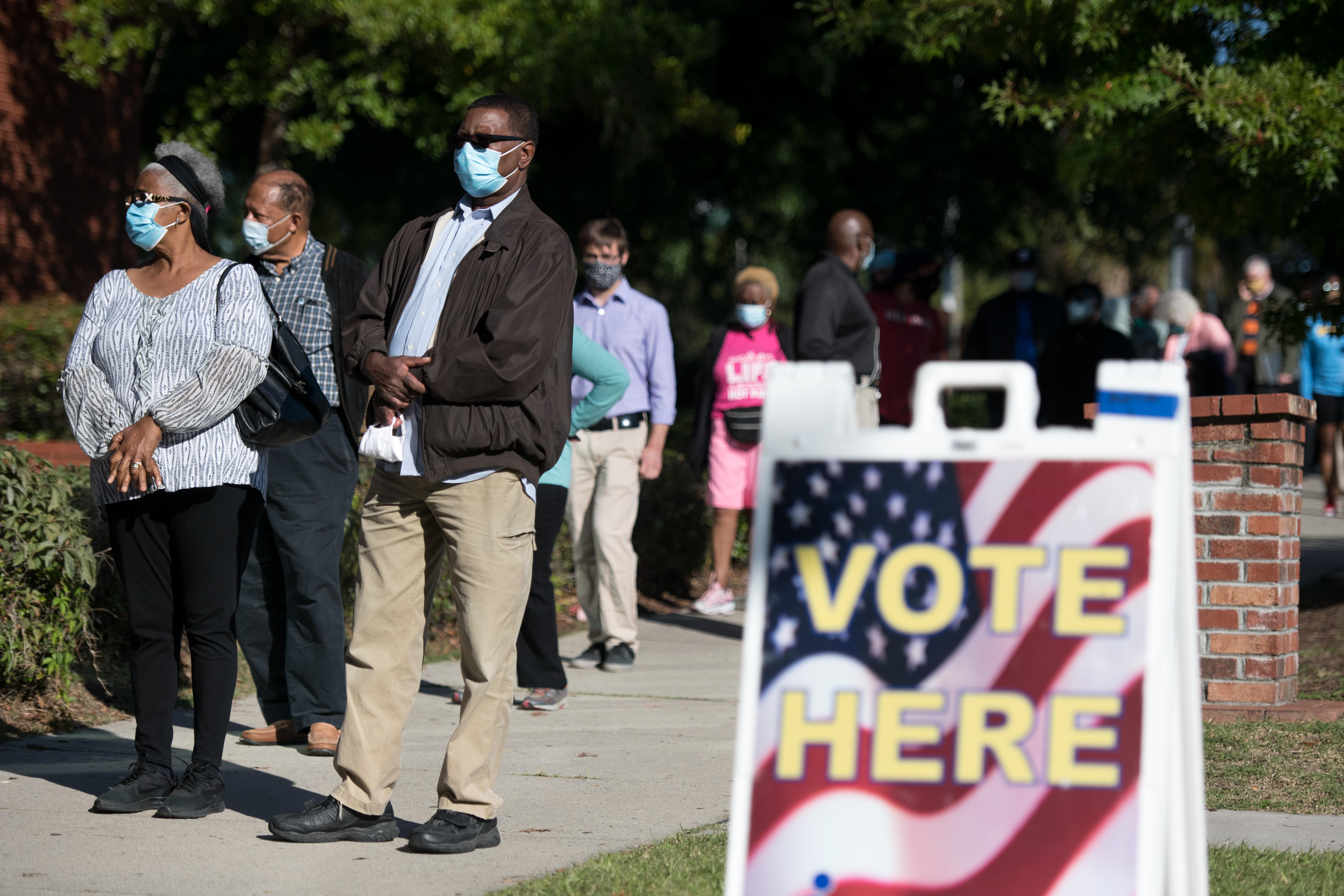 A group of voters wearing masks waits outside a polling place. A "Vote Here" sign can be seen.