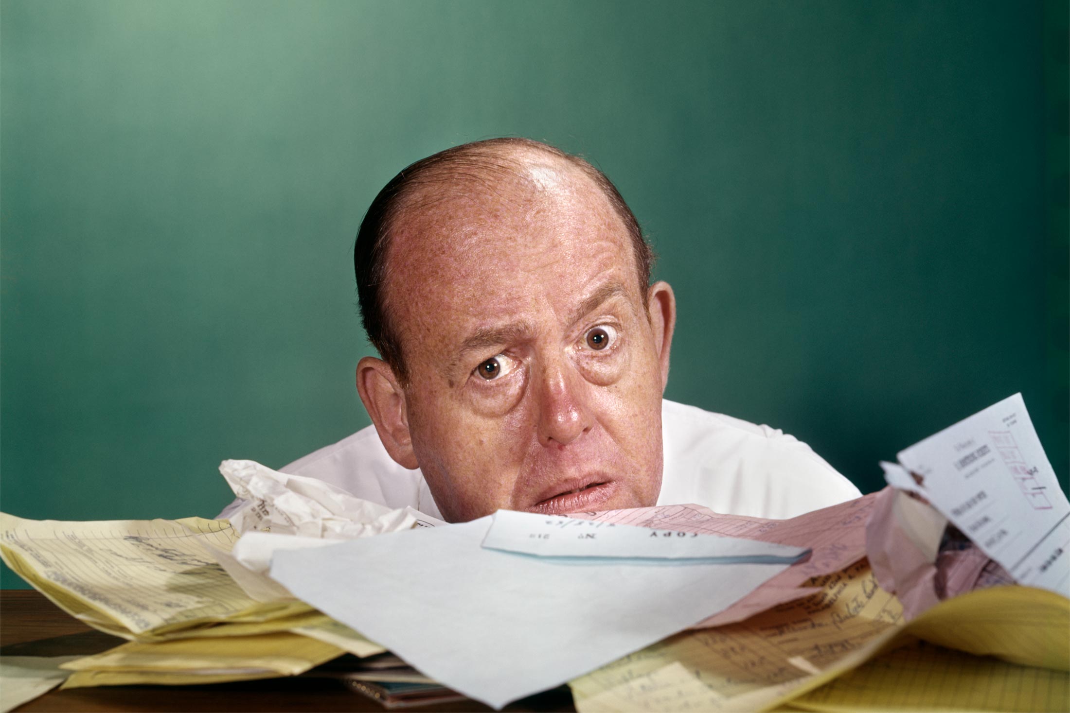 Image of a middle-aged man peering out from behind a large, messy stack of paperwork.