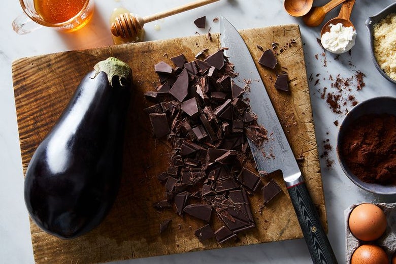 A large dark purple eggplant, a kitchen knife and lots of fragments of chocolate sit on a cutting board next to other cooking ingredients