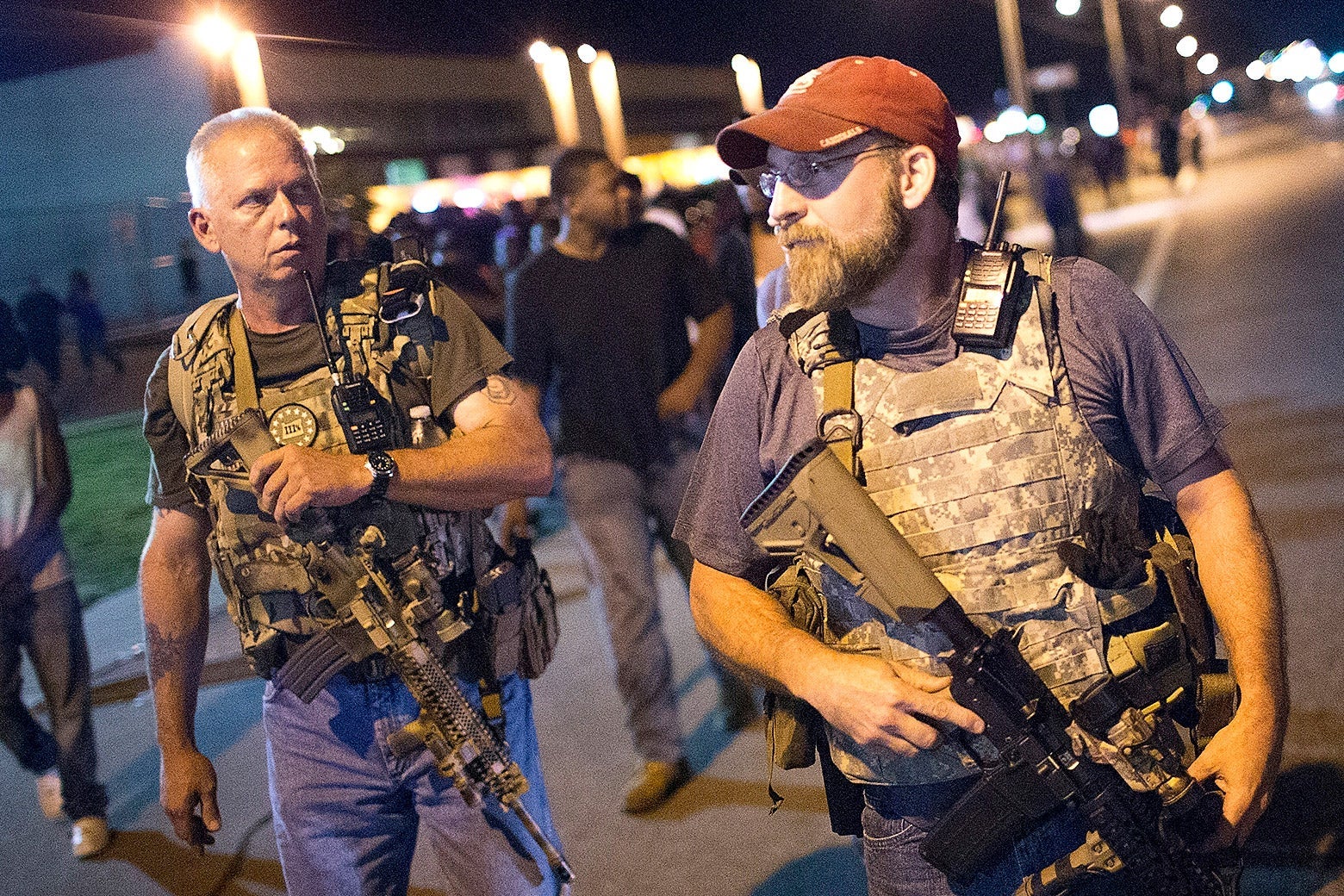 Two white Oath Keepers in tactical vests, one carrying his assault rifle and the other allowing it to hang from his holster, walk down the street. Behind them walk black protesters.