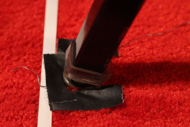 Close-up of a chair leg resting on a square of black tape next to a vertical strip of white tape on a red carpet.