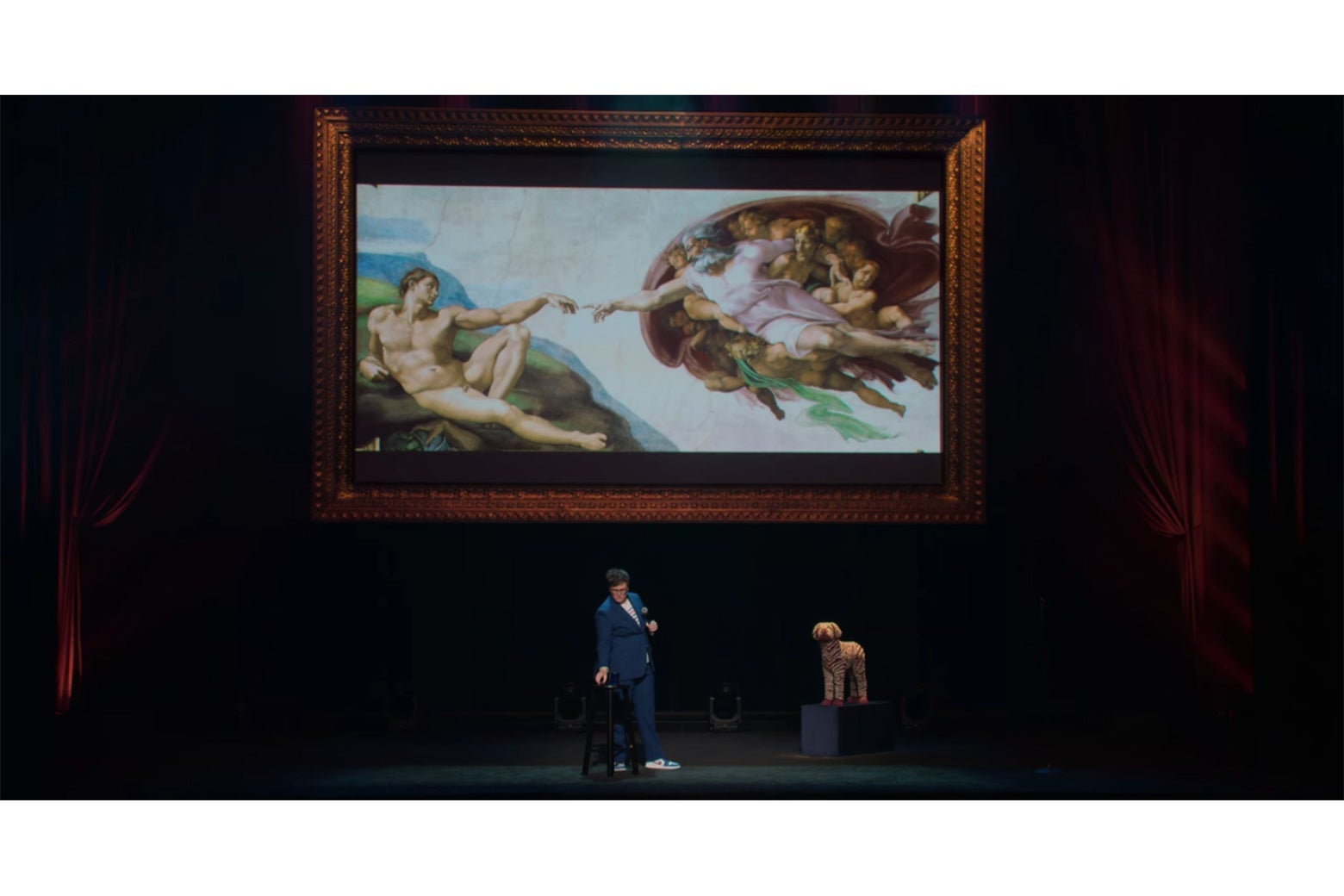 Hannah Gadsby onstage in front of a screen displaying Michelangelo's painting of God reaching out to touch Adam.