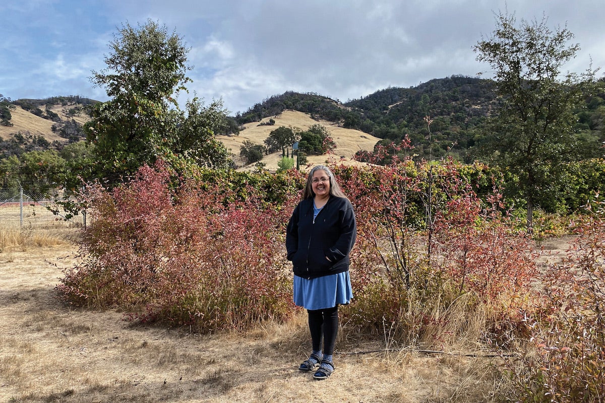 A woman stands, smiling, next to reddish bushes. In the background there are hills with pine trees and dry brush. 