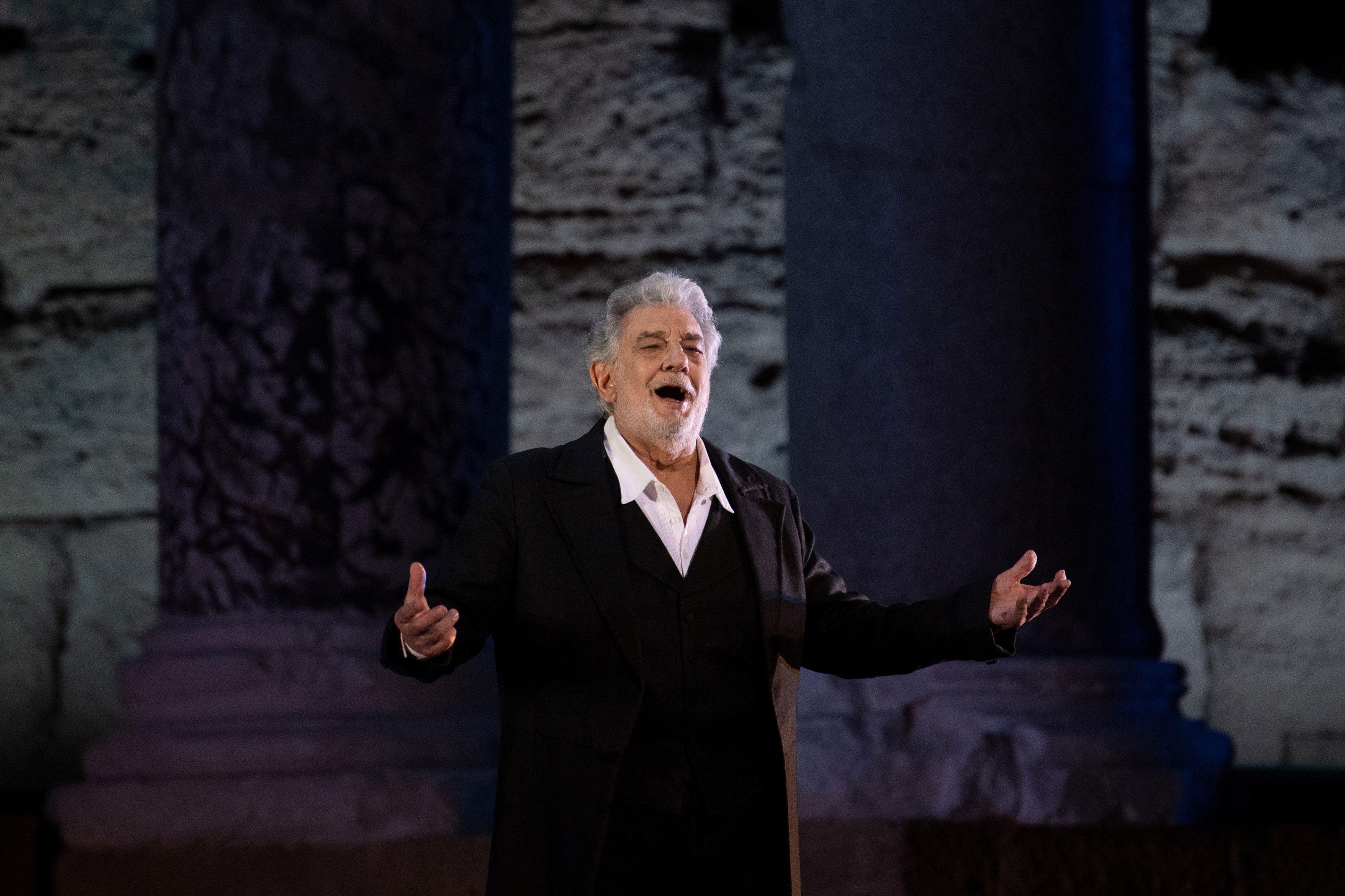 Placido Domingo stands alone on a stage, arms outstretched, mouth open.