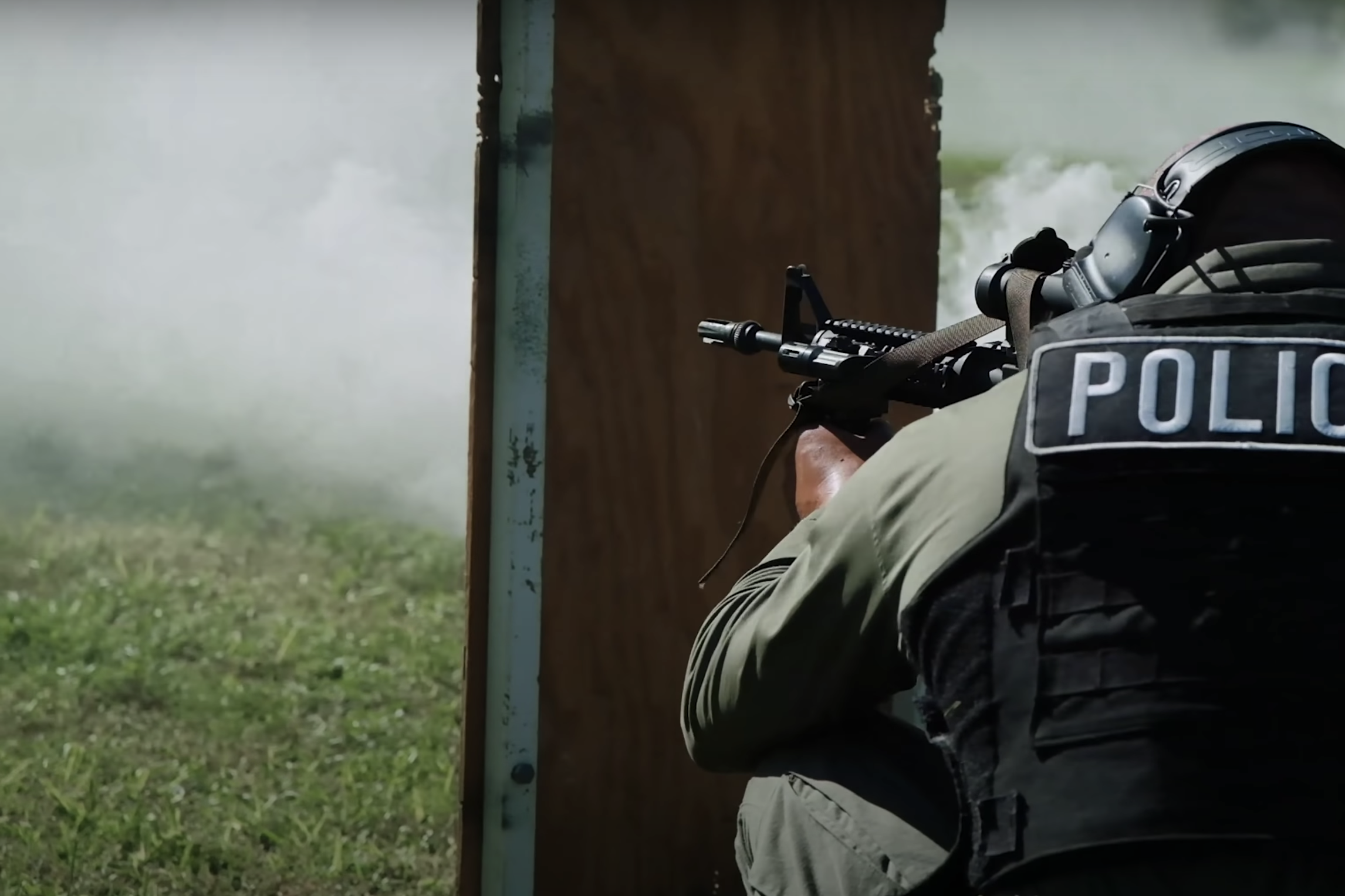 A man in a bulletproof vest holds a sniper rifle out a window.