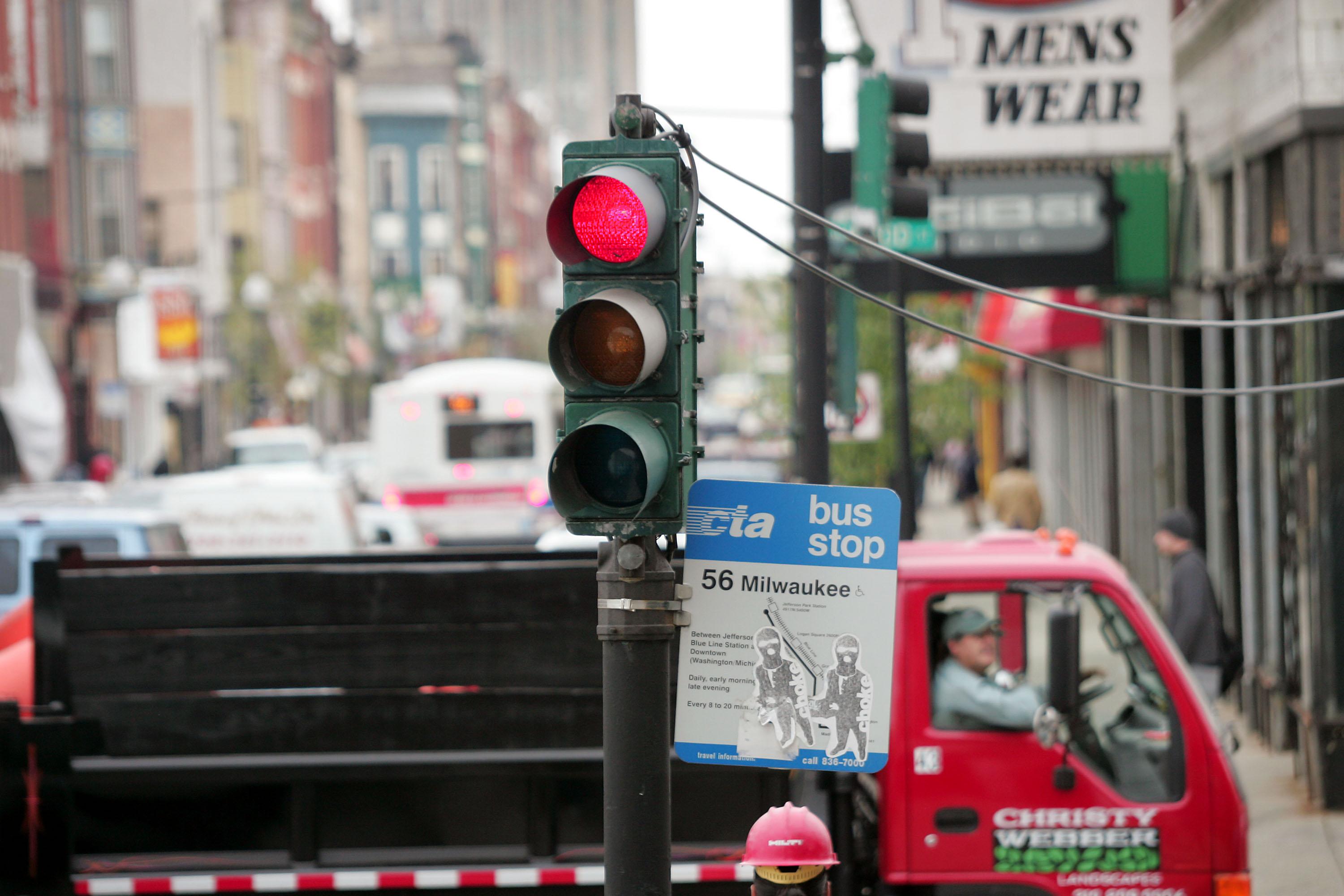 A red traffic light is in the foreground, in the background a large truck passes through a busy intersection. 
