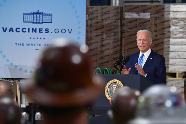 Biden gestures as he speaks at a podium to a crowd at a construction site. A vaccines.gov poster stands beside him.