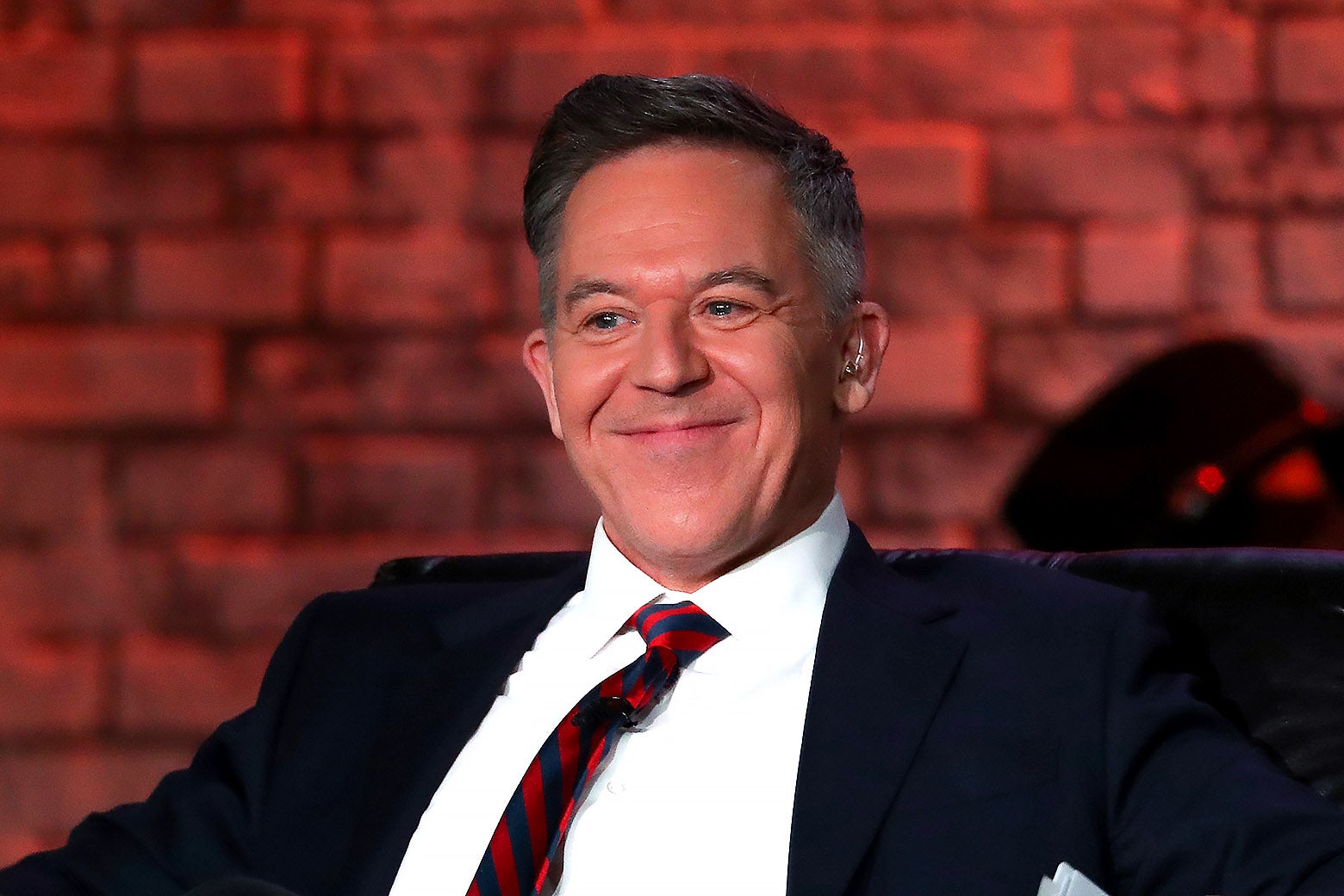 Greg Gutfeld sitting in a chair onstage with a smug grin.