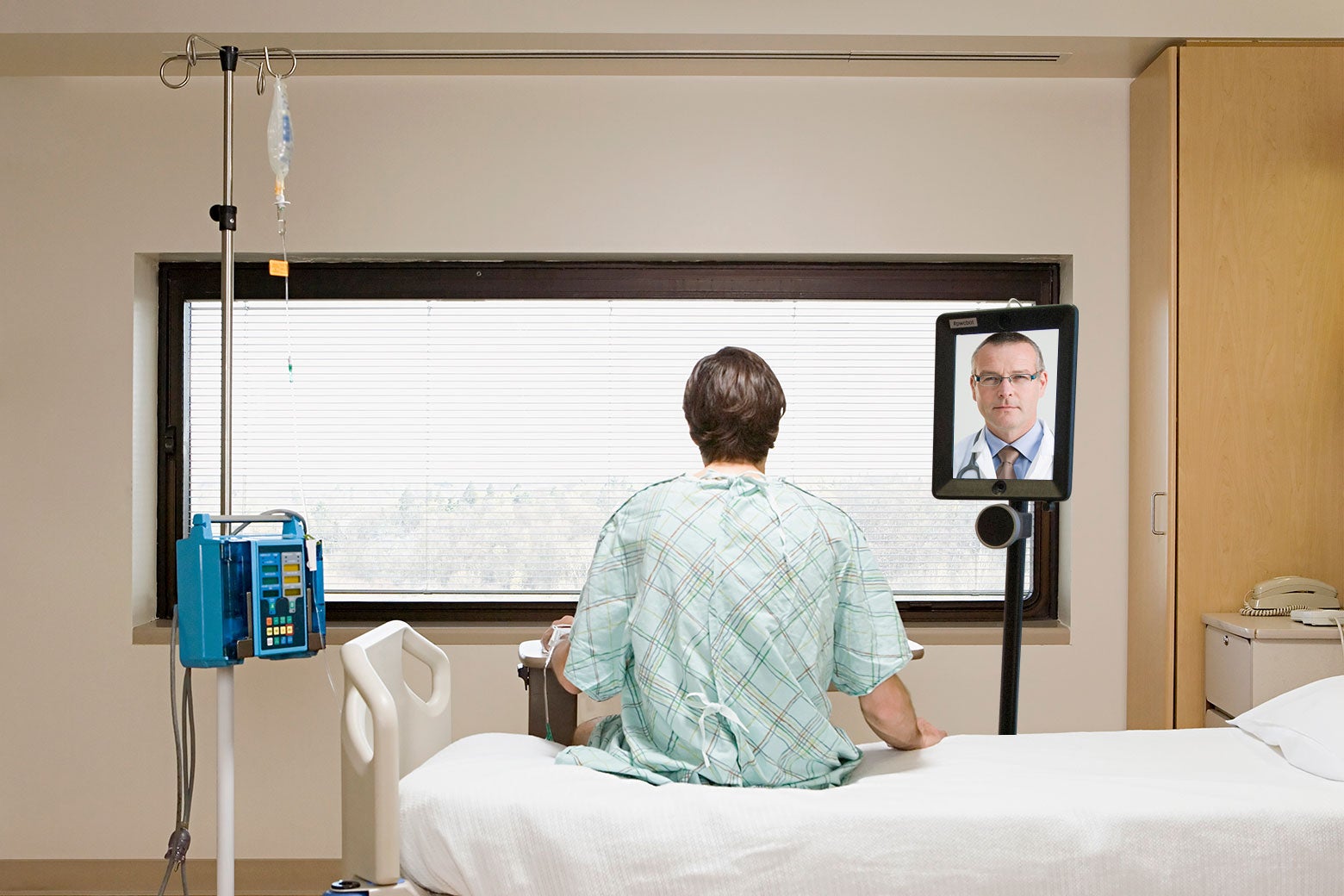 A patient sits on a hospital bed, talking to a doctor on a screen.