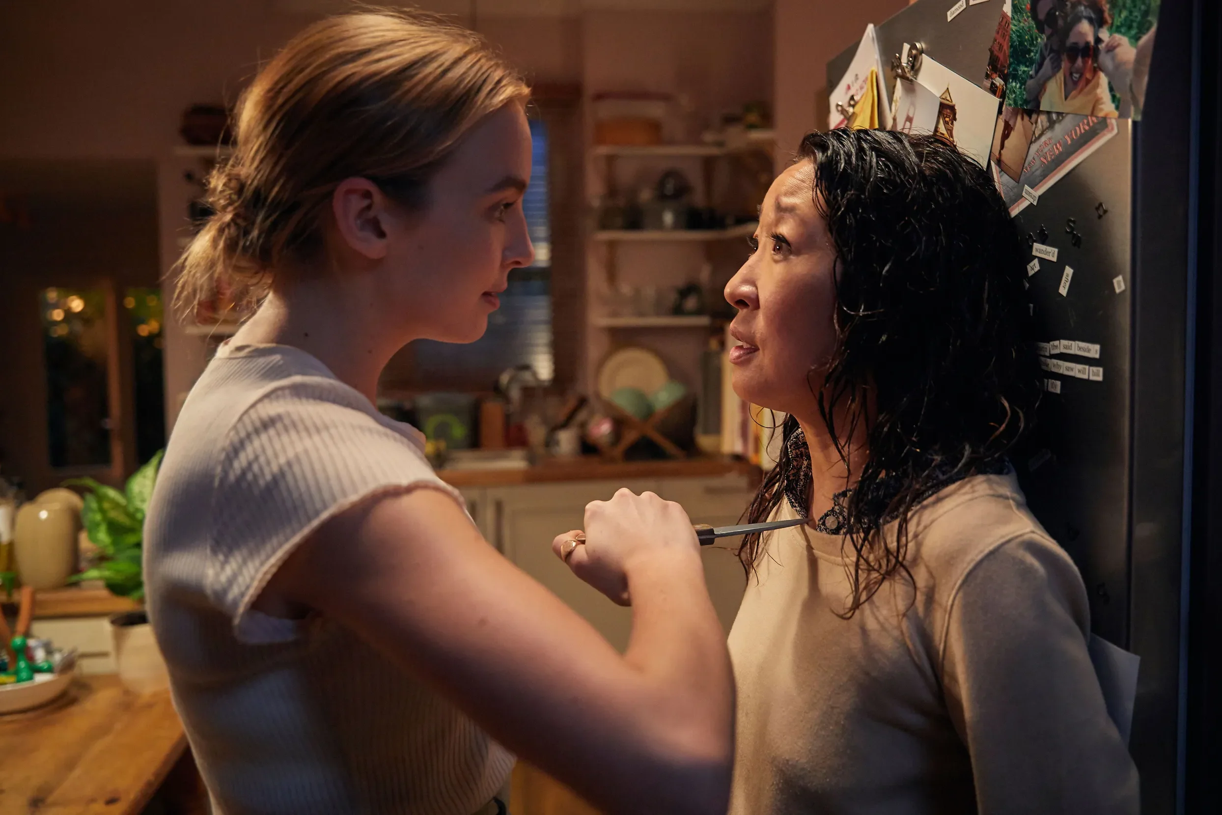 A middle-aged Asian woman stands with her back against her fridge in her homey kitchen, caught unawares by a young white woman, dressed in a white top with her hair in a bun, holding a knife to the Asian woman's neck. 