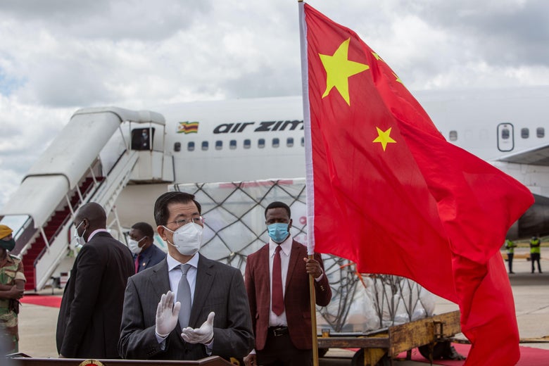 The Chinese ambassador claps with gloved hands in front of a shipment of vaccines. Behind him a Zimbabwean man holds a Chinese flag.