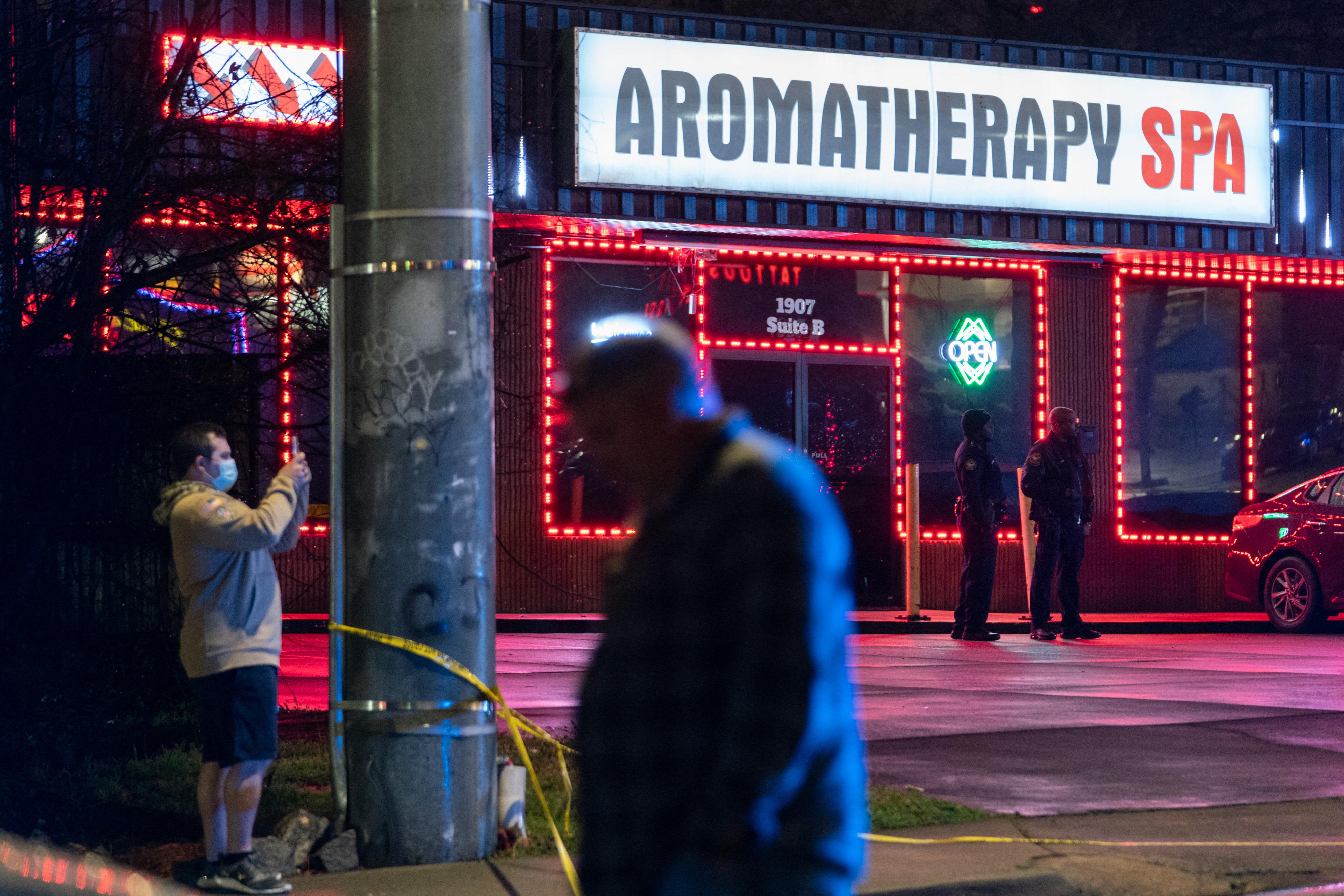 A brightly lit sign says "Aromatherapy Spa" on a building whose windows are outlined in red lights. Two law enforcement officers stand in front. It's dark outside, and a person walks in the foreground.