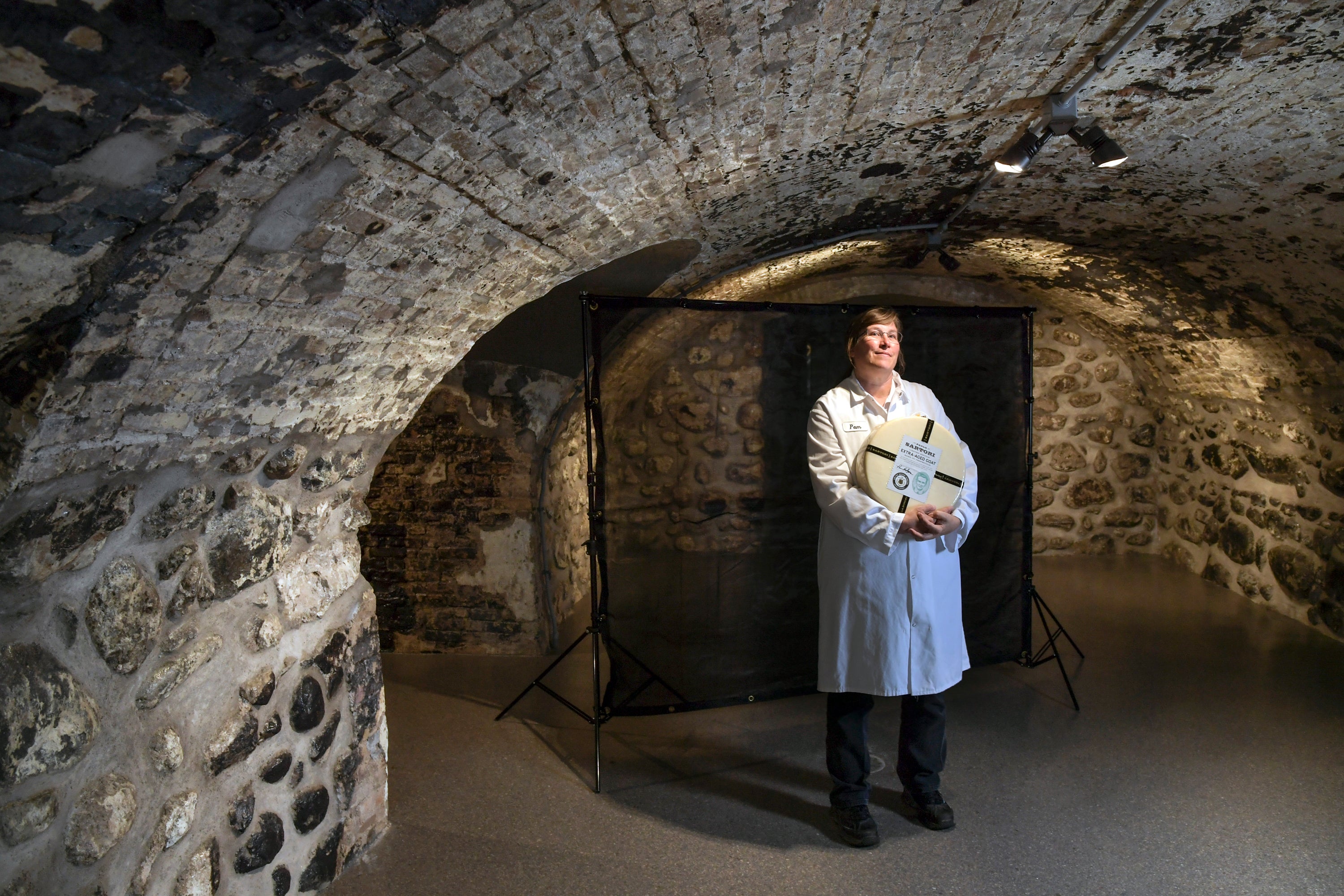 A woman holds a large wheel of cheese inside a brick-walled underground room.