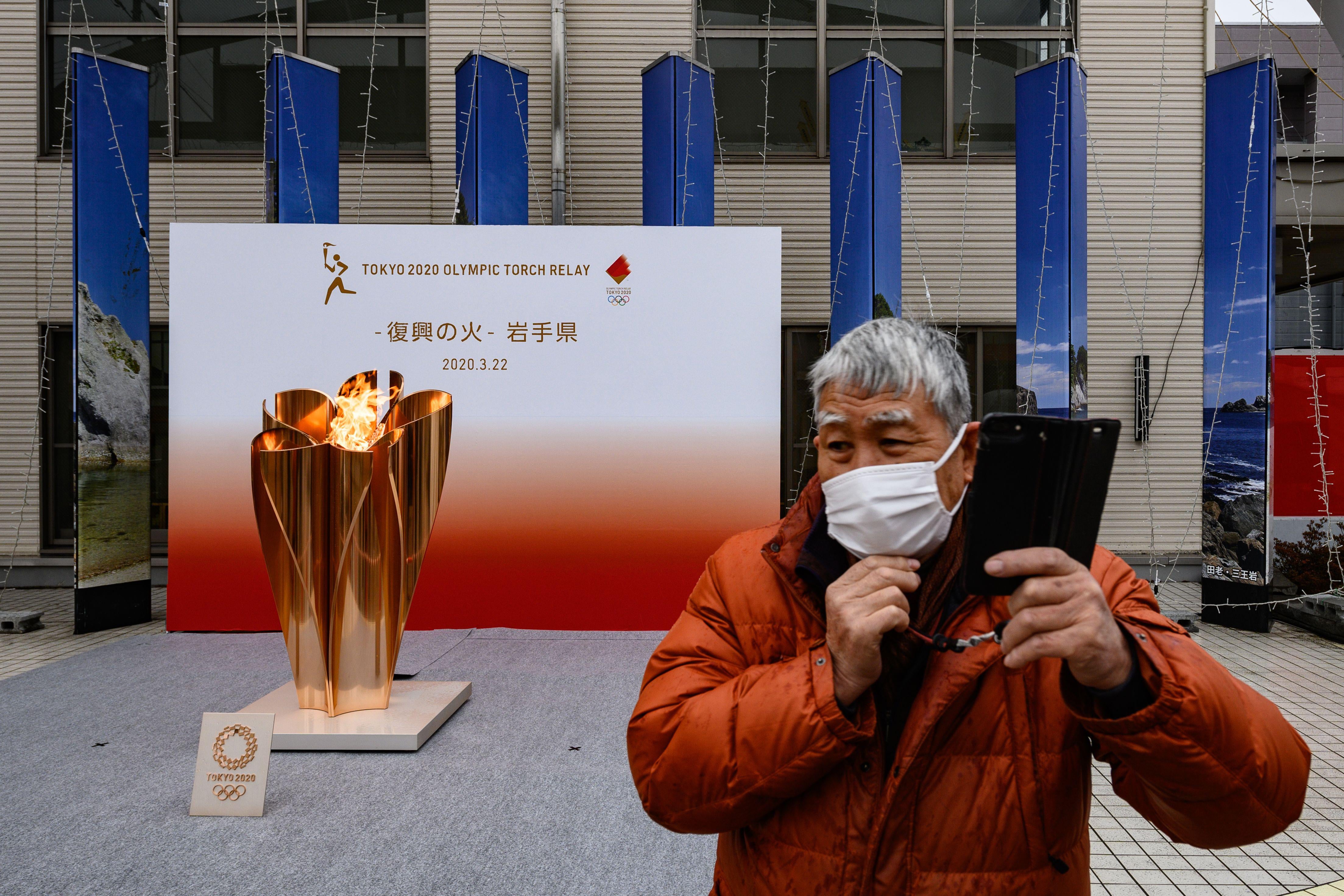 A man wearing a face mask stands in front of the Tokyo 2020 Olympic flame being displayed outside Miyako railway station, Iwate prefecture on March 22, 2020, after arriving from Greece.