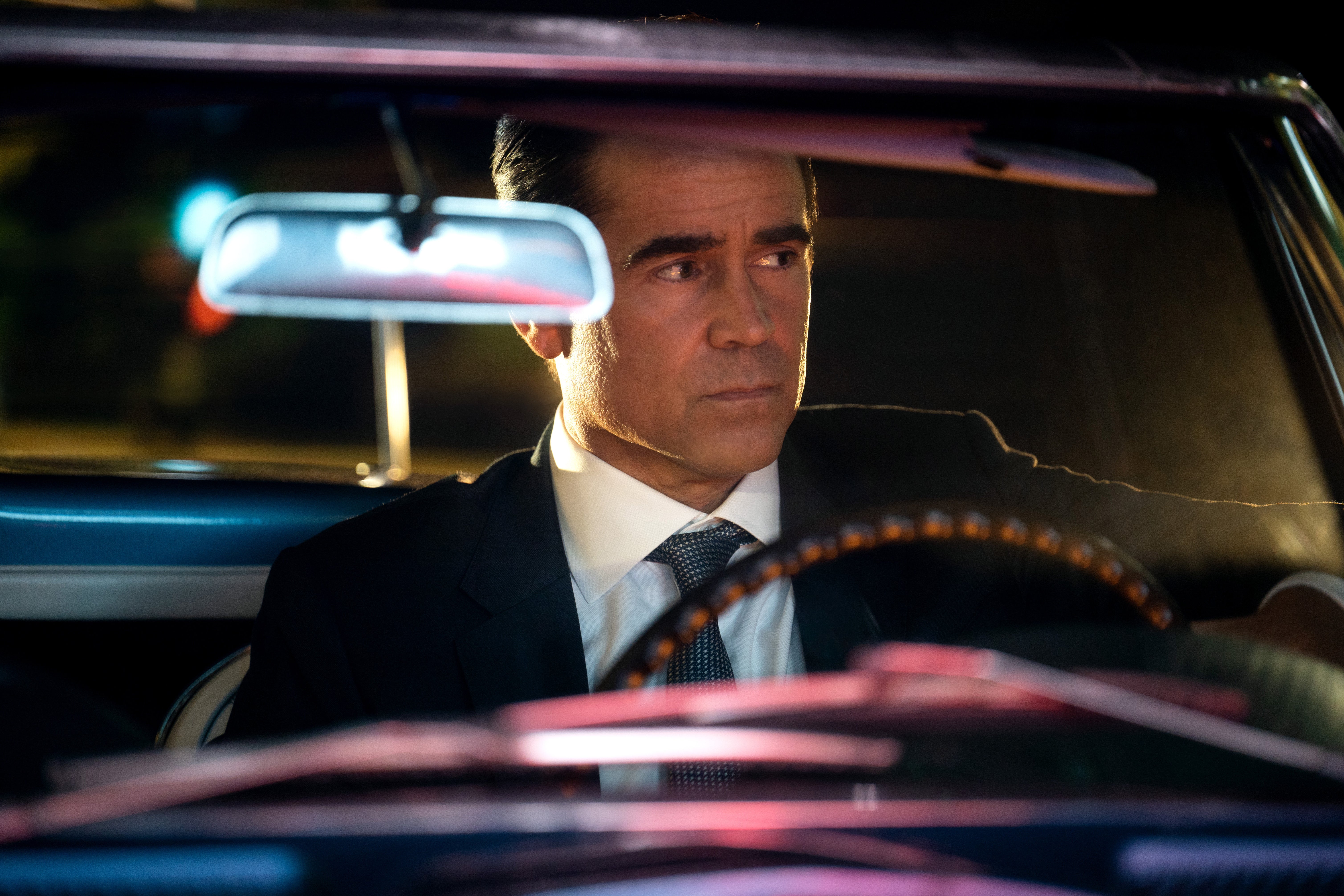 Colin Farrell as Detective Sugar, wearing a suit and driving at night, is lit by neon lights as he looks off to the side. 