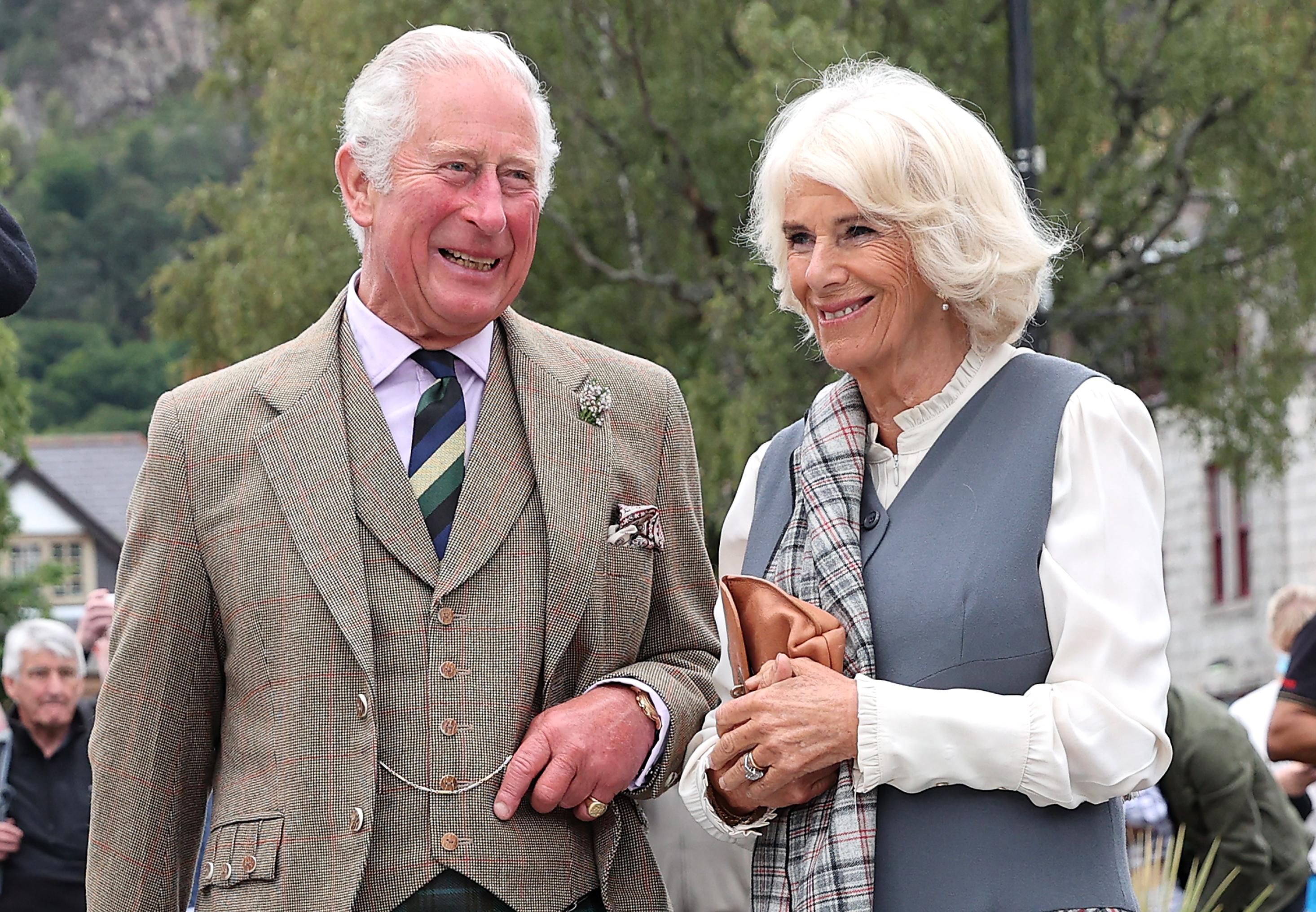 Charles and Camilla smile while standing side by side. He is wearing a plaid three-piece suit and she is wearing a grey vest, white blouse, and plaid scarf. 